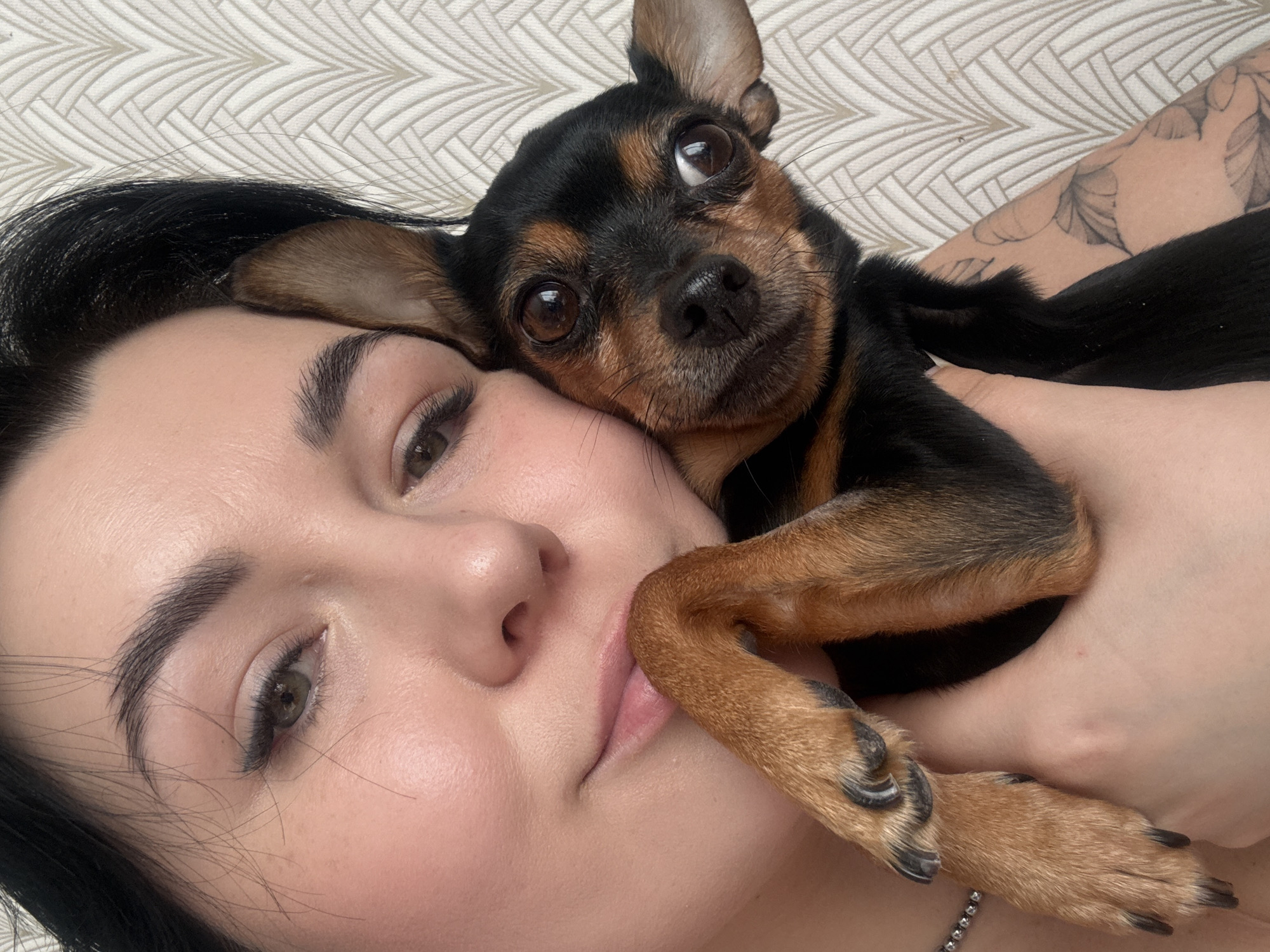 An indoor close-up selfie, potentially from Chaikovsky, Russia, captures a tender moment between a person and a small dog. The person, appearing to be female with dark hair, defined eyebrows, and subtle eye makeup, is seen lying down with a relaxed expression and slightly parted lips. A part of a silver-colored necklace or chain is visible on their neck.
The small black and tan dog, possibly a Miniature Pinscher or a similar toy breed, is nestled affectionately against the person's face, resting its head on their cheek and neck. The dog's large, round brown eyes are wide and attentive, looking towards the camera. Its front paws are gently placed on the person's arm and chest, with the person's hand subtly holding the dog.
A delicate tattoo with a floral or leafy pattern is visible on the person's arm. The background features a light-colored, geometrically patterned surface, suggesting a headboard, pillow, or patterned wall covering. The lighting is soft and even, indicating an indoor setting but not a specific time of day. The overall mood is one of warmth, affection, and companionship between the human and their pet. No text is visible in the image.