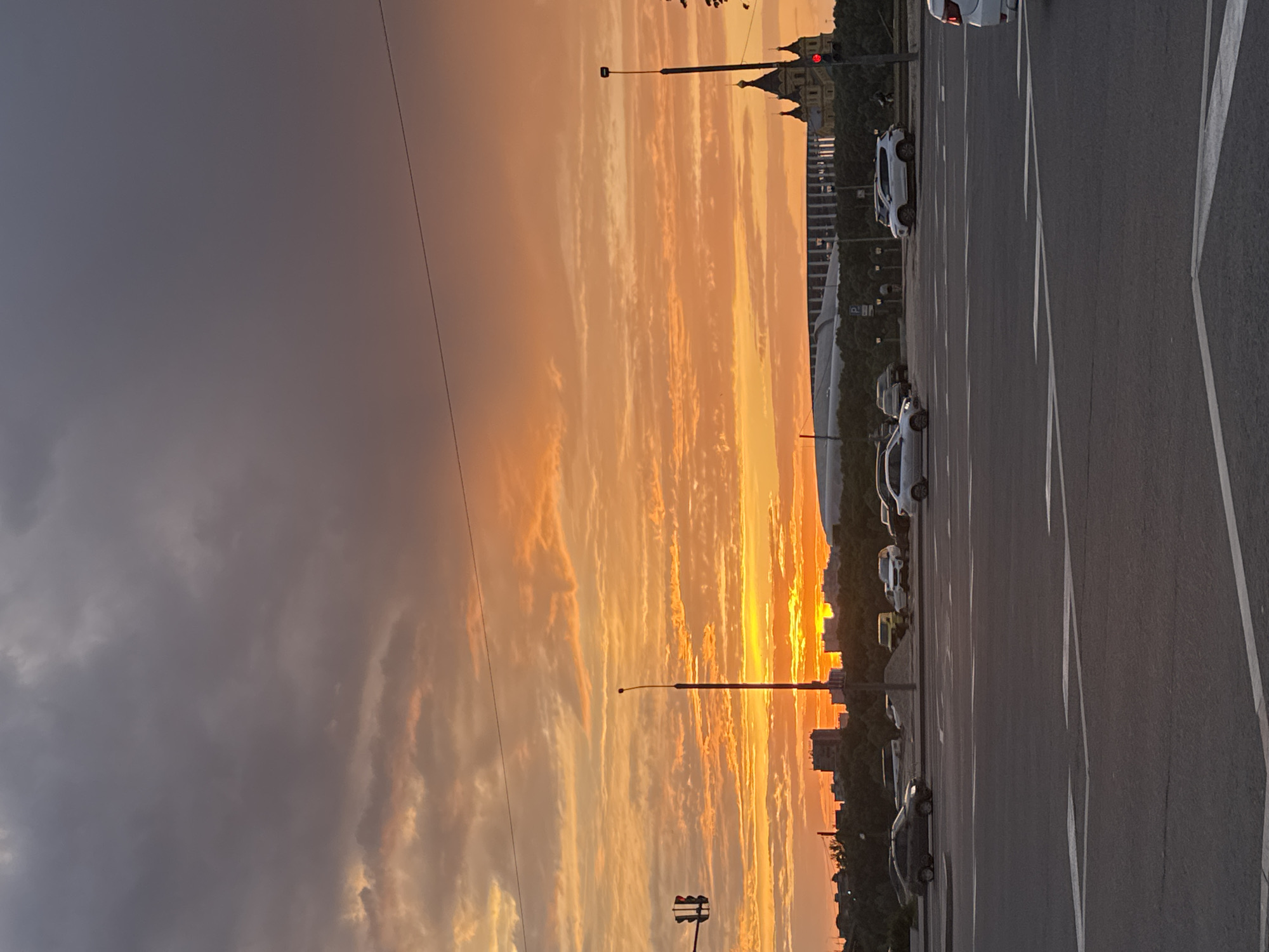 The image shows a sunset view from a parking area in Nizhny Novgorod, Russia, likely near the Alexander Nevsky Cathedral. In the foreground, there is a paved parking lot with several parked cars along the right edge of the frame. There are multiple white parking lines visible on the dark asphalt. Streetlights punctuate the view towards the horizon. In the background, the skyline is silhouetted against a vivid sunset, displaying a gradient from bright yellow and orange near the horizon to softer orange and grey hues higher in the sky. The sky features layers of clouds. On the right side of the horizon, a silhouette of the Alexander Nevsky Cathedral and other buildings is visible. A traffic light is located in the lower left.
