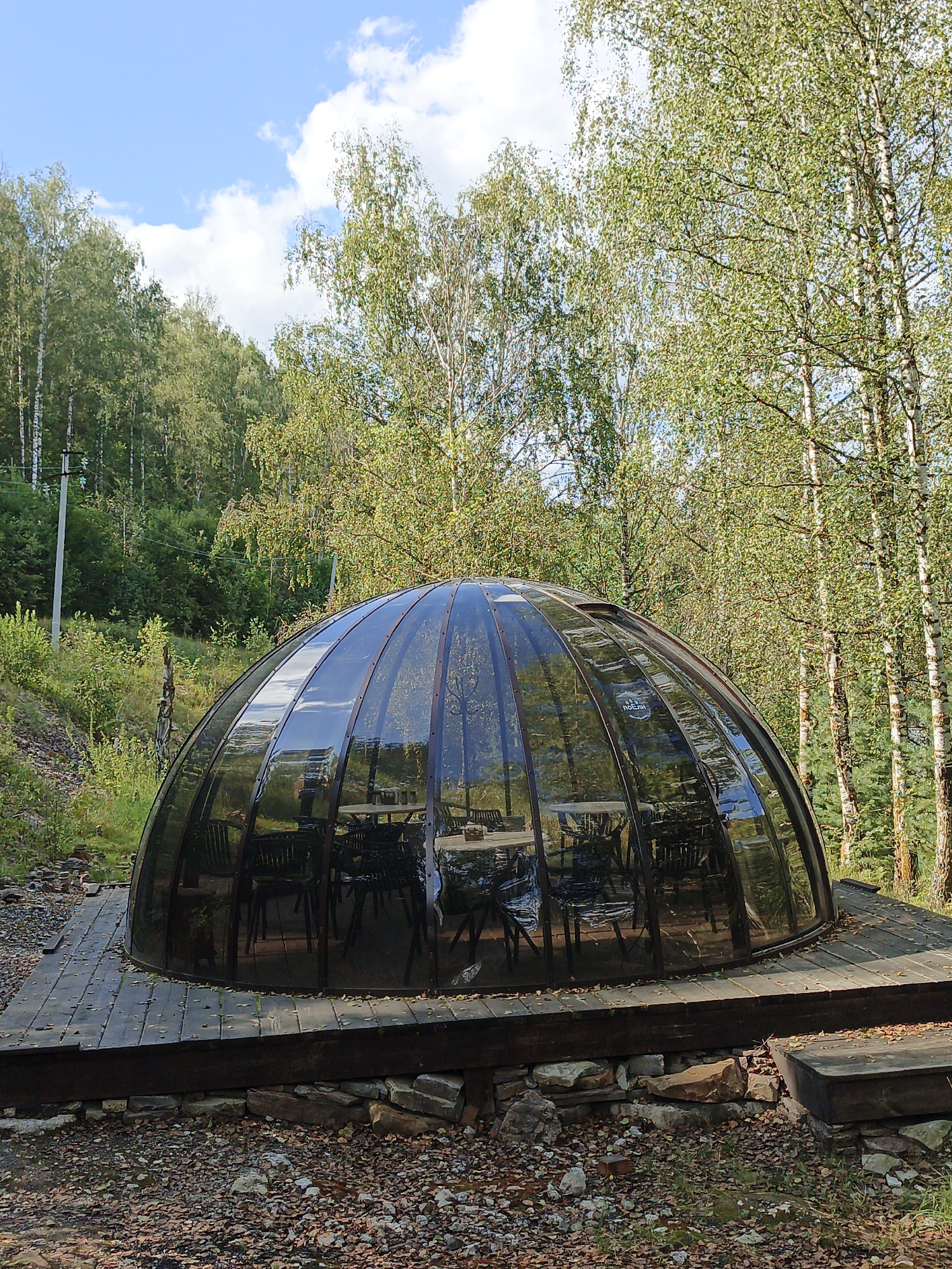 A geodesic dome structure with dark-tinted glass panels sits on a wooden platform. Inside the dome, several tables and chairs are visible. The structure is surrounded by trees, with birch trees being prominent in the background. The ground is covered with gravel and fallen leaves. The scene appears to be in an outdoor, natural setting. The specific location is the Porogi village in Russia, an area known for its hydroelectric power station and surrounding natural beauty. There are no people visible in the image.