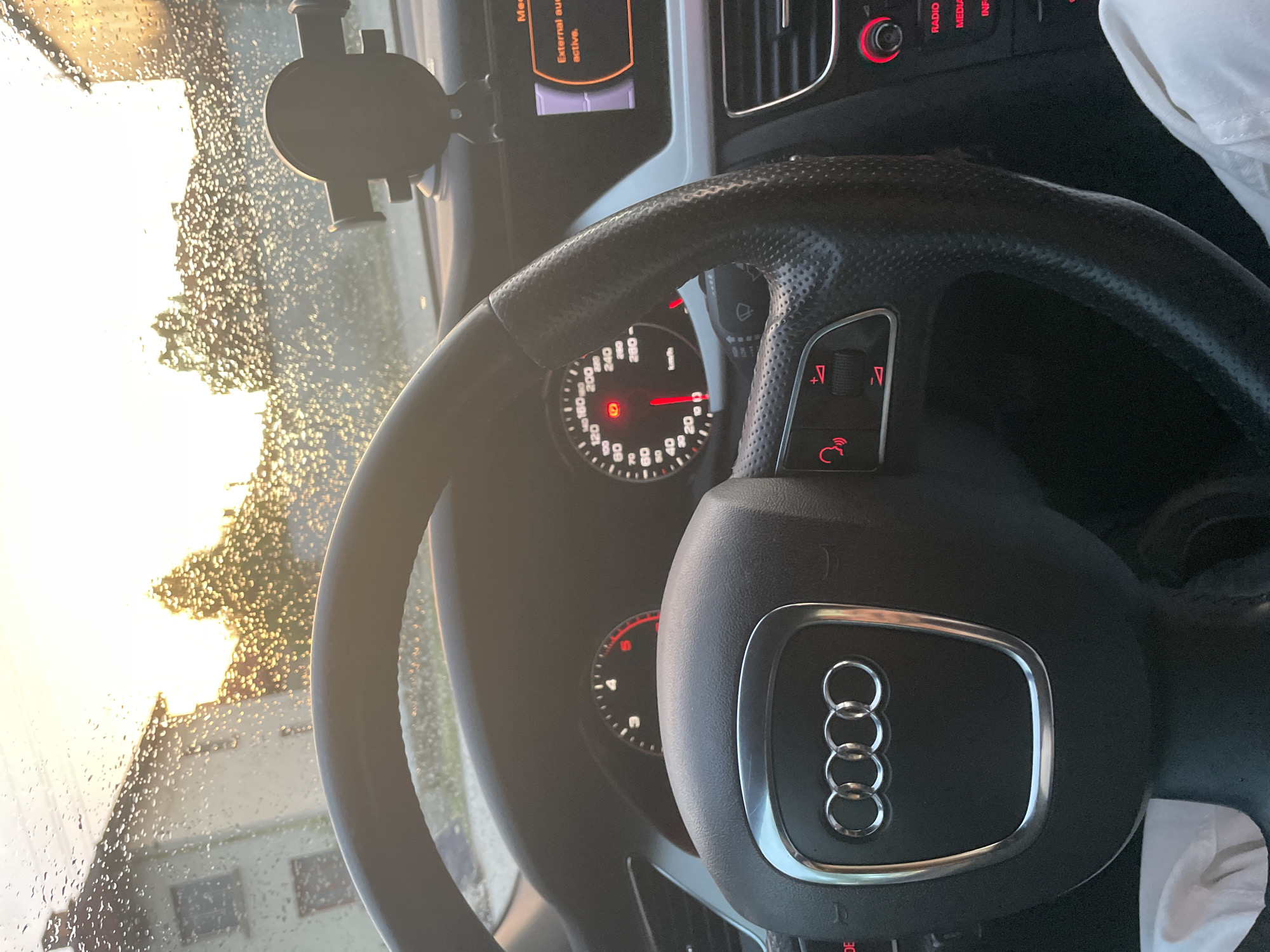 Inside the driver's seat of an Audi vehicle in Ihtiman, Bulgaria, the image captures the dashboard, steering wheel, and a view through a rain-splattered windshield. The prominent black leather steering wheel features the Audi four-ring logo at its center and multi-function buttons, including volume controls (indicated by "V+" and "V-") and a call icon. Behind the steering wheel, the instrument cluster displays a speedometer with markings up to "280" and a tachometer showing RPMs up to "6," with a red warning light (possibly a parking brake indicator) illuminated. To the right, various car controls such as "RADIO," "MEDIA," and "INFO" buttons are visible.
A digital screen on the dashboard shows the message "External audio active." A phone holder is mounted on the windshield, which is covered in numerous raindrops. Beyond the windshield, bright, golden sunlight penetrates through the water droplets, illuminating blurred outlines of buildings or trees, suggesting either sunrise or sunset. The overall scene implies the car is stationary or moving slowly in rainy weather during the golden hour. A white piece of clothing or fabric is visible on the passenger side.
