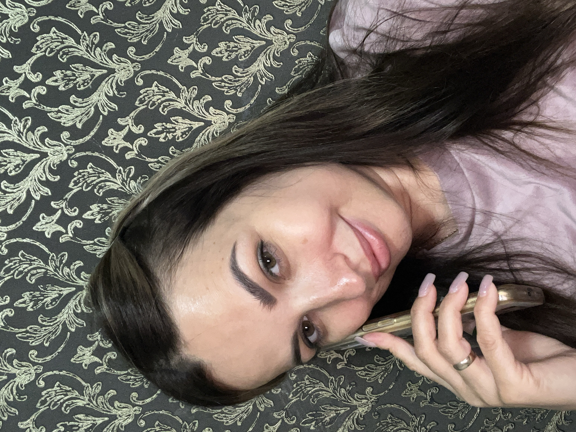 In a close-up shot, a woman with dark hair and brown eyes is holding a gold-colored smartphone to her ear. She is wearing a pink shirt and a gold ring on her left ring finger. The background is a dark wallpaper with a gold damask pattern. The woman is smiling. The setting is indoors.