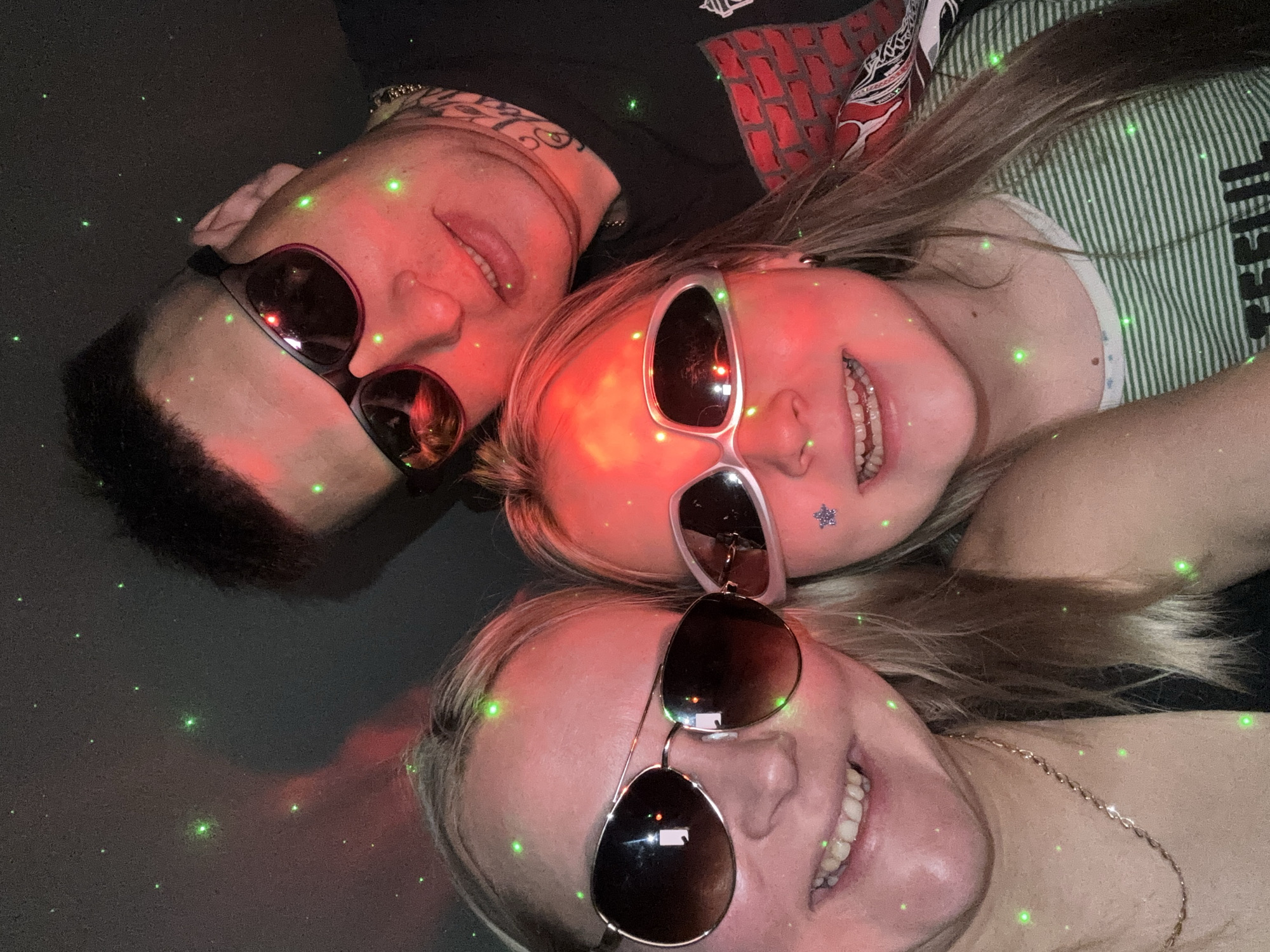 Three people are lying down, facing upwards, and wearing sunglasses. The individual at the top left has dark hair and tattoos visible on their face and neck. The person in the middle is wearing pink sunglasses and has a star sticker on their cheek. The person at the bottom is wearing sunglasses with brown lenses. The environment appears to be indoors, with a dark background illuminated by green laser-like dots and red lighting. The location of Izhevsk, Russia is not evident.