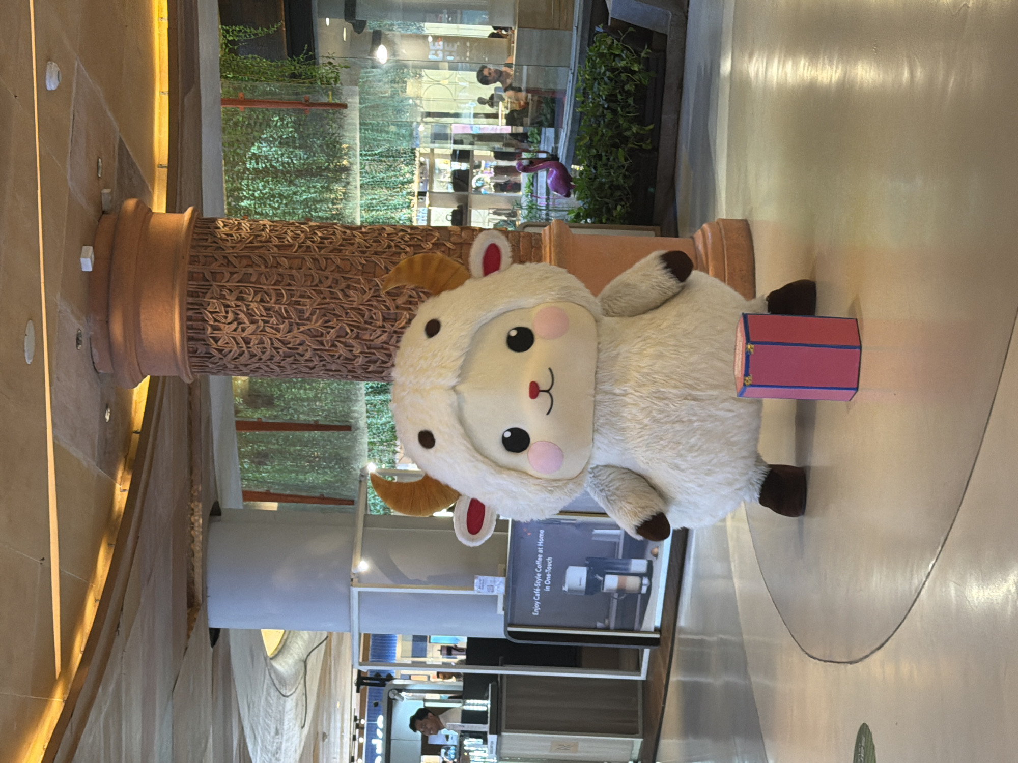 The image shows a large stuffed sheep toy positioned in a shopping center, likely in Kuta, Indonesia. The sheep is fluffy and white with brown accents. It is placed next to a textured brown pillar. In the background, there is a storefront and plants. The floor is light-colored and shiny. In the foreground, there's a small pink box. The shop is likely a food and beverage cafe. A man and some food is visible behind the glass facade.