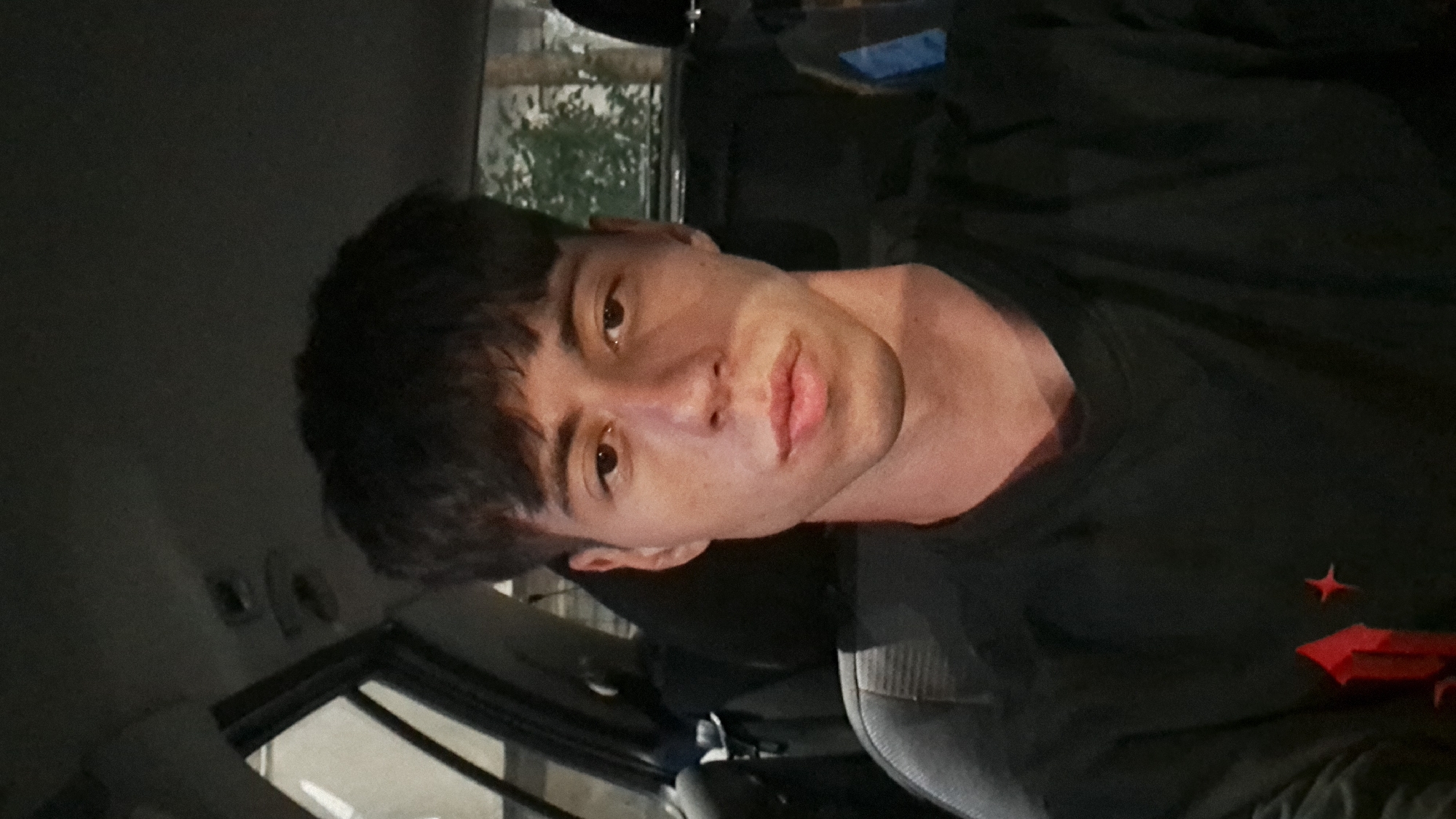 A close-up, frontal shot shows a young man with dark, tousled hair and light skin, wearing a black t-shirt with a red emblem on the left side of the chest. He is seated in the driver's seat of a car. The background is dark and out of focus, with glimpses of what appears to be foliage and parts of the car's interior. The lighting suggests it is nighttime. No specific details about the location in Burgas, Bulgaria are visible in the image.