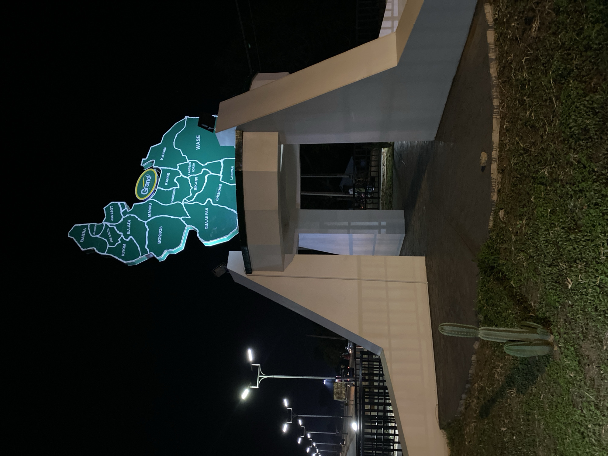 This is a night-time, medium-shot, eye-level, outdoor photograph of a monument in Bukuru, Nigeria. The monument is an illuminated green map of Plateau State, Nigeria, with names of local government areas labeled within it. A yellow oval with the word "Grand!" is superimposed on the map. The map is mounted on a white concrete structure that forms an archway. In the foreground, to the left of the archway, are two cacti plants. Beyond the archway, through the opening, streetlights and vehicles are visible in the distance. The lighting in the image is artificial, emanating from the monument and the streetlights. The overall scene depicts a public landmark at night.