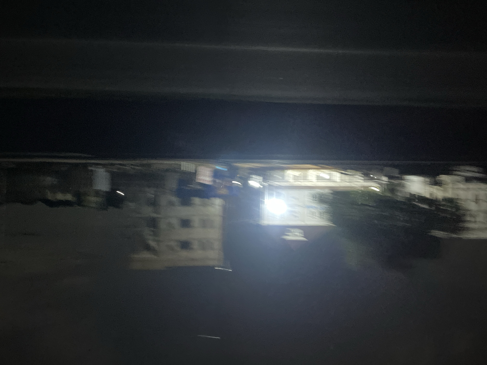 At night in Gwarinpa, Nigeria, a blurred and dark cityscape is visible. Buildings are illuminated by bright lights, while trees can also be seen.