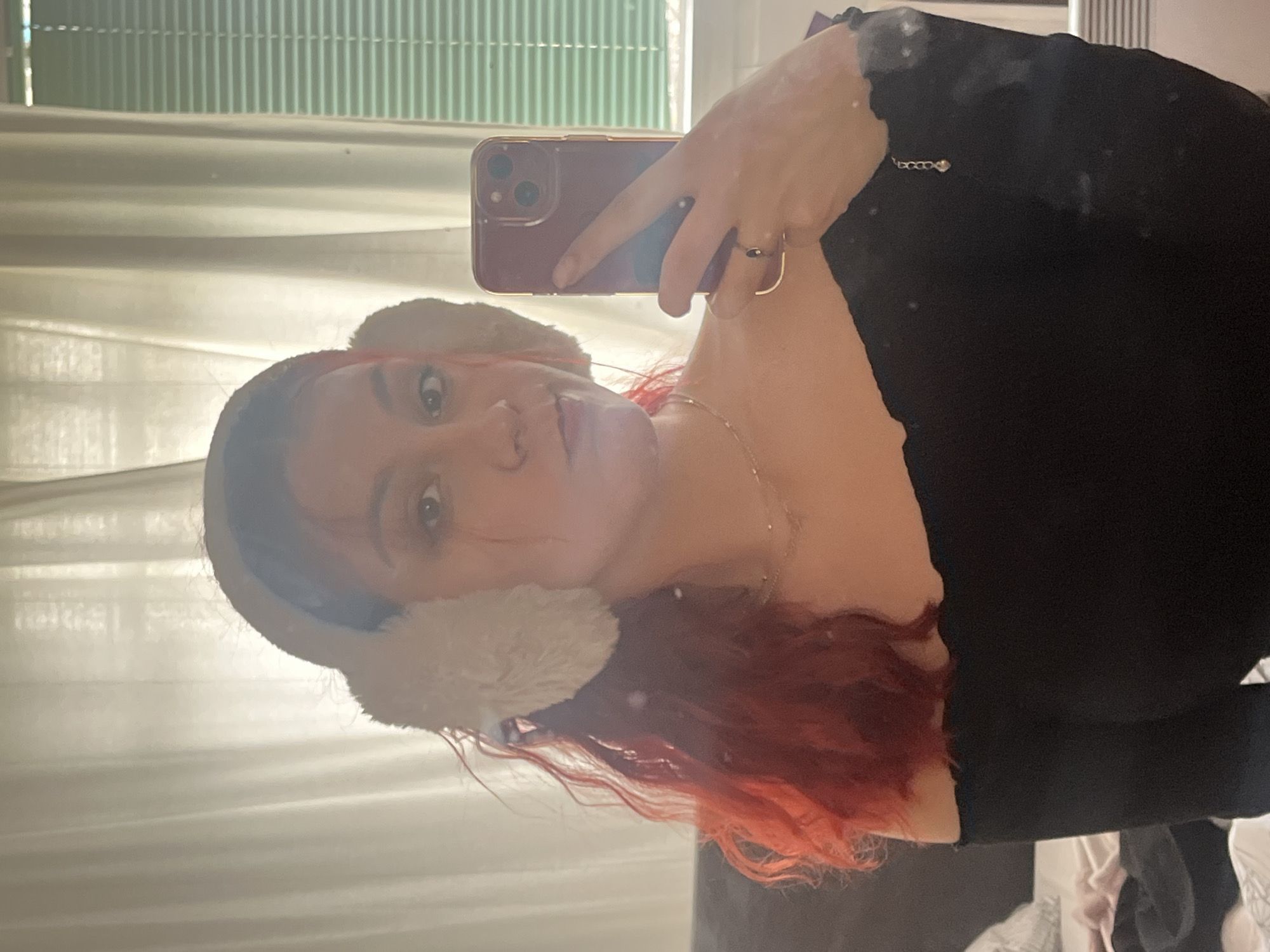 A young woman with red hair is taking a selfie in a room with curtains and a green venetian blind in the background. She is wearing beige earmuffs and a black shirt. She is holding a red smartphone in her hand, which has a ring on her finger. She is wearing a necklace. The location is inside an apartment, and it is not possible to determine if it is located in Sofia, Bulgaria.