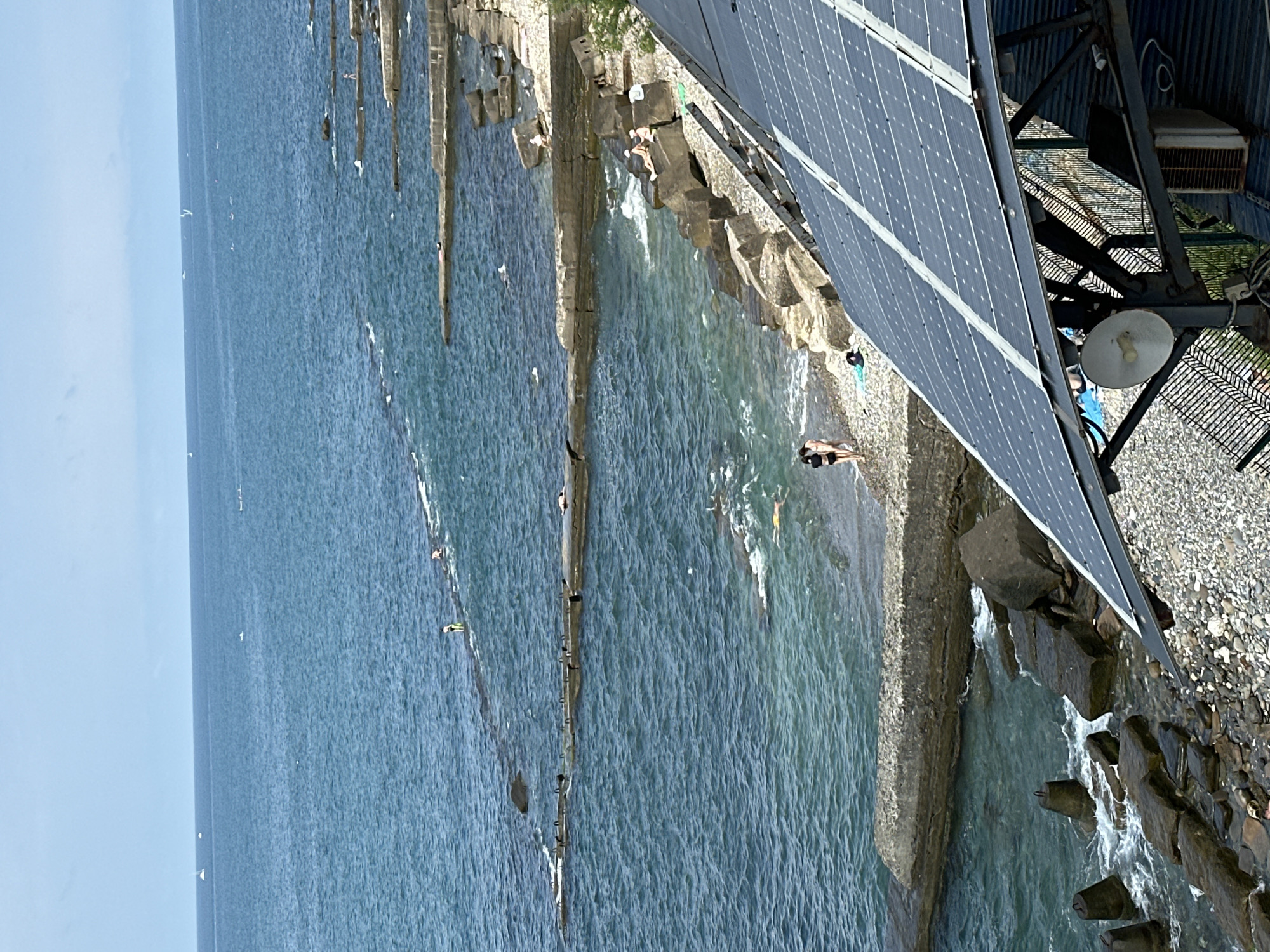 This image captures a view of the Black Sea coast in Sochi, Russia, on a clear day. In the foreground, a portion of a solar panel installation is visible, angled downwards and to the right, resting on a metal frame. Beyond the solar panels, a pebbled beach transitions into a rocky coastline. Several large, gray rocks are scattered along the shore, with some extending into the water.
The sea is a vibrant blue, with visible waves gently lapping against the shore and the constructed breakwaters. These breakwaters, made of concrete and stone, extend out into the water in a series of parallel lines. Several people are present along the coastline and in the water. On the beach, two individuals in swimsuits are walking. Further out, along the breakwaters, other individuals are seen sunbathing or wading in the shallow water. In the distance, several sailboats and small boats are visible on the horizon. The sky is a pale blue, with some lighter cloud cover.