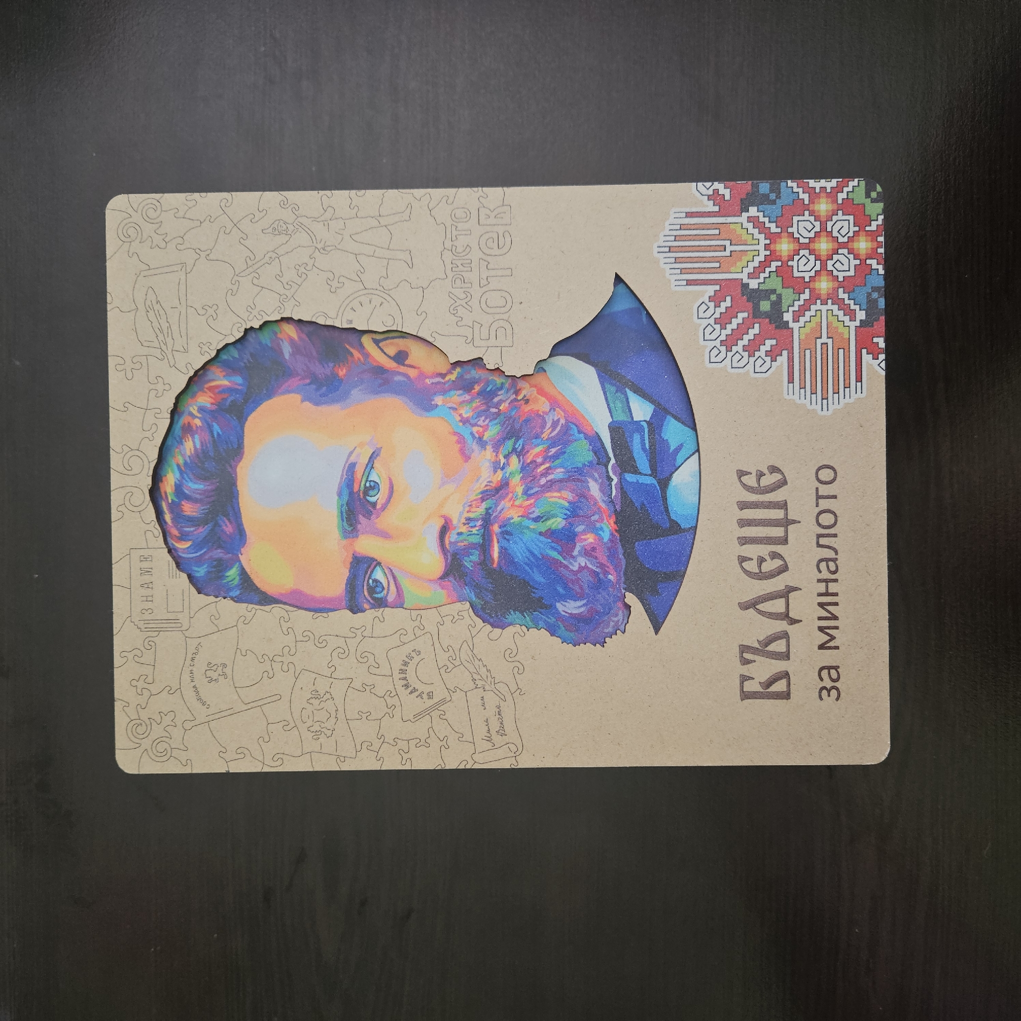 The image shows a rectangular object, likely a book or notebook cover, lying on a dark, flat surface. The cover features a stylized, colorful portrait of a man with a beard and mustache. The portrait is cut out of the cover material, revealing a jigsaw puzzle-like pattern underneath. Above the portrait is a decorative, pixelated design with red, green, blue, yellow, and white colors. To the right of the portrait, in Bulgarian Cyrillic script, are the words "БЪДЕЩЕ за миналото" (Bǎdešče za minaloto), which translates to "Future for the past." Scattered around the man's portrait and incorporated into the jigsaw puzzle pattern are small drawings of items such as a feather quill, a clock, a flag, and other objects. There is the phrase "Христо Ботев" near the cut-out image. The objects and text suggest the subject of the cover is related to history, possibly Bulgarian.