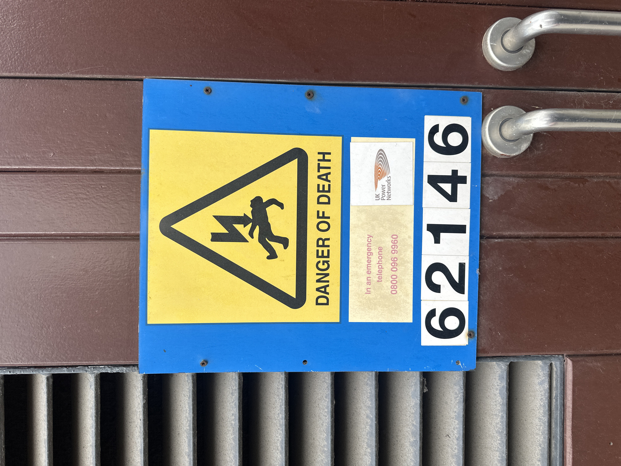 The image shows a blue danger sign attached to a brown, corrugated metal structure, likely an electrical substation, and is located in London, United Kingdom. The sign prominently displays a yellow triangle with a black silhouette of a person being struck by lightning, accompanied by the words "DANGER OF DEATH" in black lettering. Adjacent to this is a smaller rectangular area with the logo for "UK Power Networks" and contact information, including the emergency telephone number 0800 096 9960. To the right of the sign are white numbers "62146". The sign is affixed to the structure with screws. Beneath the sign, there is a metal grate. Above the sign, there are two handles made of metal, and the doors they are attached to, are closed.