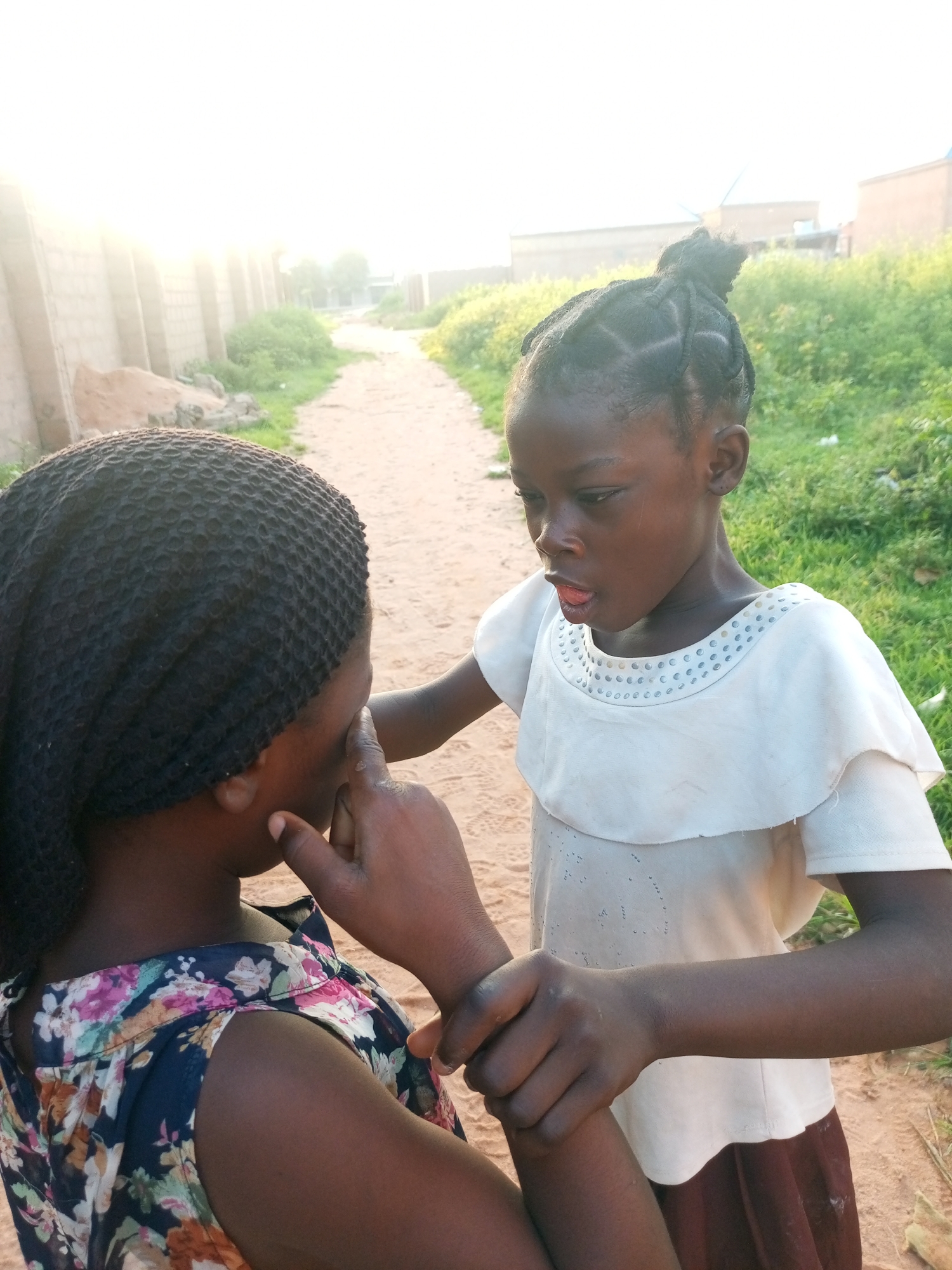 In Tempure, Nigeria, a young Black girl with braided hair and a white shirt is seen touching the face of another Black girl who is wearing a floral print top. The interaction takes place outdoors on a dirt path, with green grass and vegetation lining the path. In the background, there are buildings and a wall constructed of concrete blocks, with the bright sun creating a strong glare.