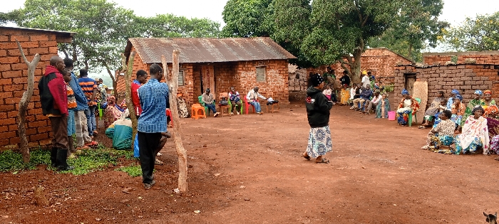In an outdoor village setting, a gathering of men and women are grouped around an open dirt area. Some people are seated on chairs or the ground while others are standing, engaging in conversation. Buildings constructed from red bricks and with tin roofs are scattered in the background. Trees provide shade, creating a communal space for social interaction. A woman holding a child walks through the open space towards the seated group.