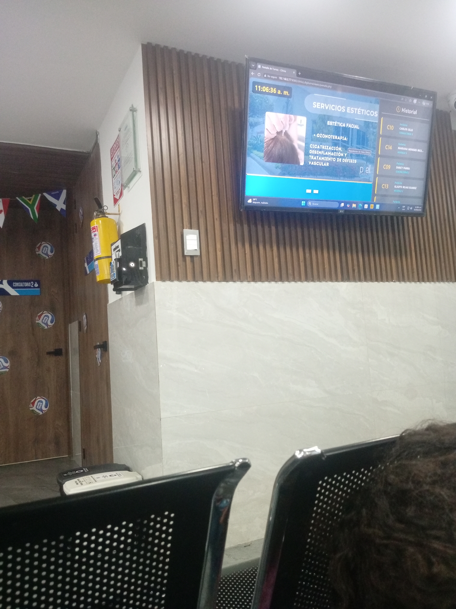 The image shows an interior waiting area, likely a clinic or medical office, in Perímetro Urbano Cúcuta, Colombia. In the background, a flat-screen television displays a digital queue system for "SERVICIOS ESTÉTICOS." The screen shows the time as "11:06:36 a. m." and lists various aesthetic treatments such as "ESTÉTICA FACIAL" and "OZONOTERAPIA." Patient names and corresponding clinic room numbers (C10, C14, C09, C13) are visible.
To the left of the television, a yellow fire extinguisher is mounted on a wall. Several small flags, including the South African flag, are pinned to the wall. Further to the left, a blue sign reads "CONSULTORIO 2." Decorative soccer balls are also affixed to the wooden paneling on the left wall.
In the foreground, two black perforated chairs are partially visible. The right chair is occupied by a person whose head and dark, curly hair are visible. The environment appears to be clean and well-lit, with light-colored tiled walls. The overall impression is that of a modern, organized waiting space.