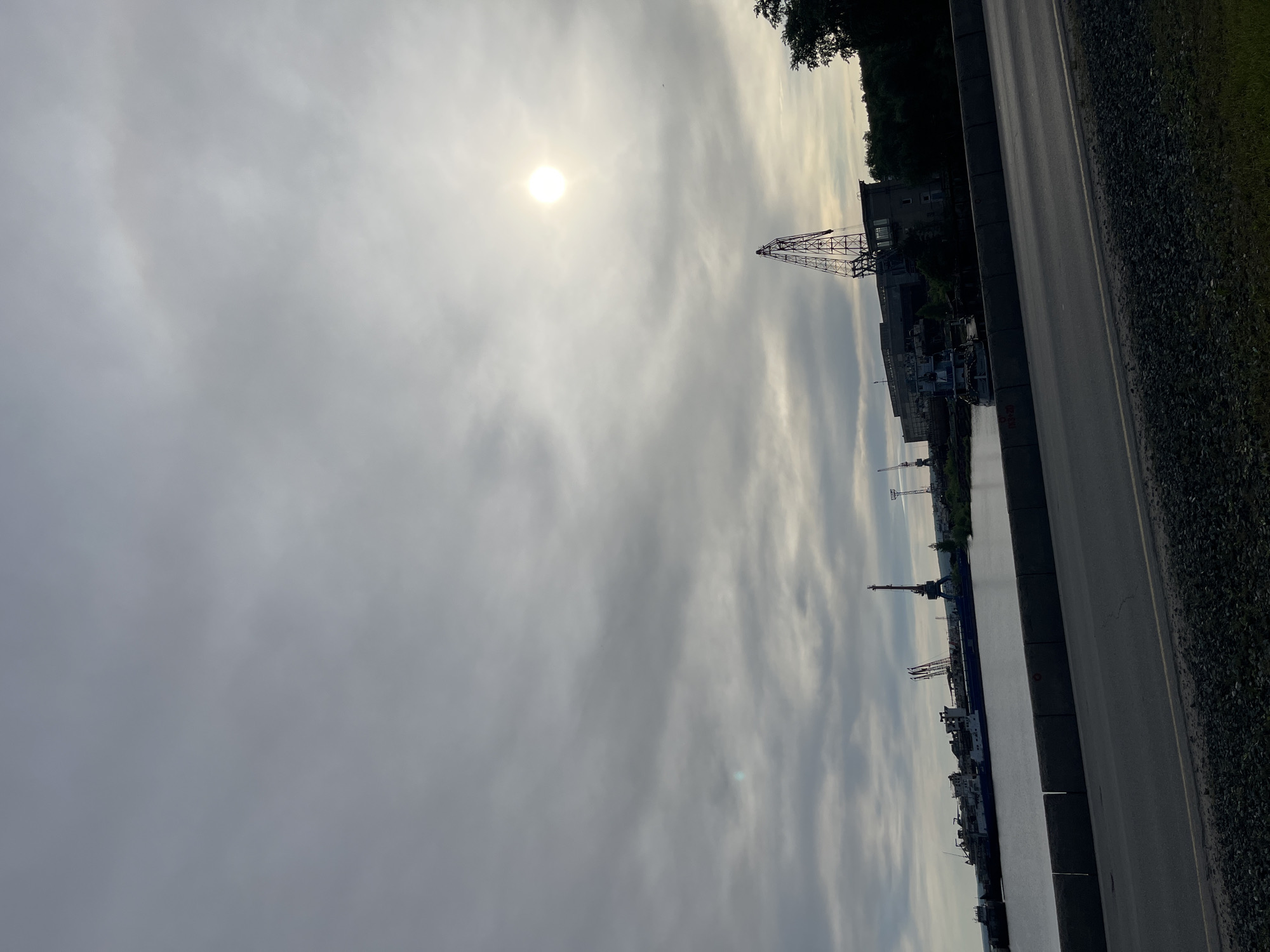 The image shows a view along the embankment in the city of Chaikovsky, Russia. The sky is overcast with the sun visible through the clouds. Along the embankment is a road with a guardrail. Beyond the road is the river, in which several ships are docked. Cranes are visible on the ships and along the riverbank. The buildings of the port area and vegetation are also visible.