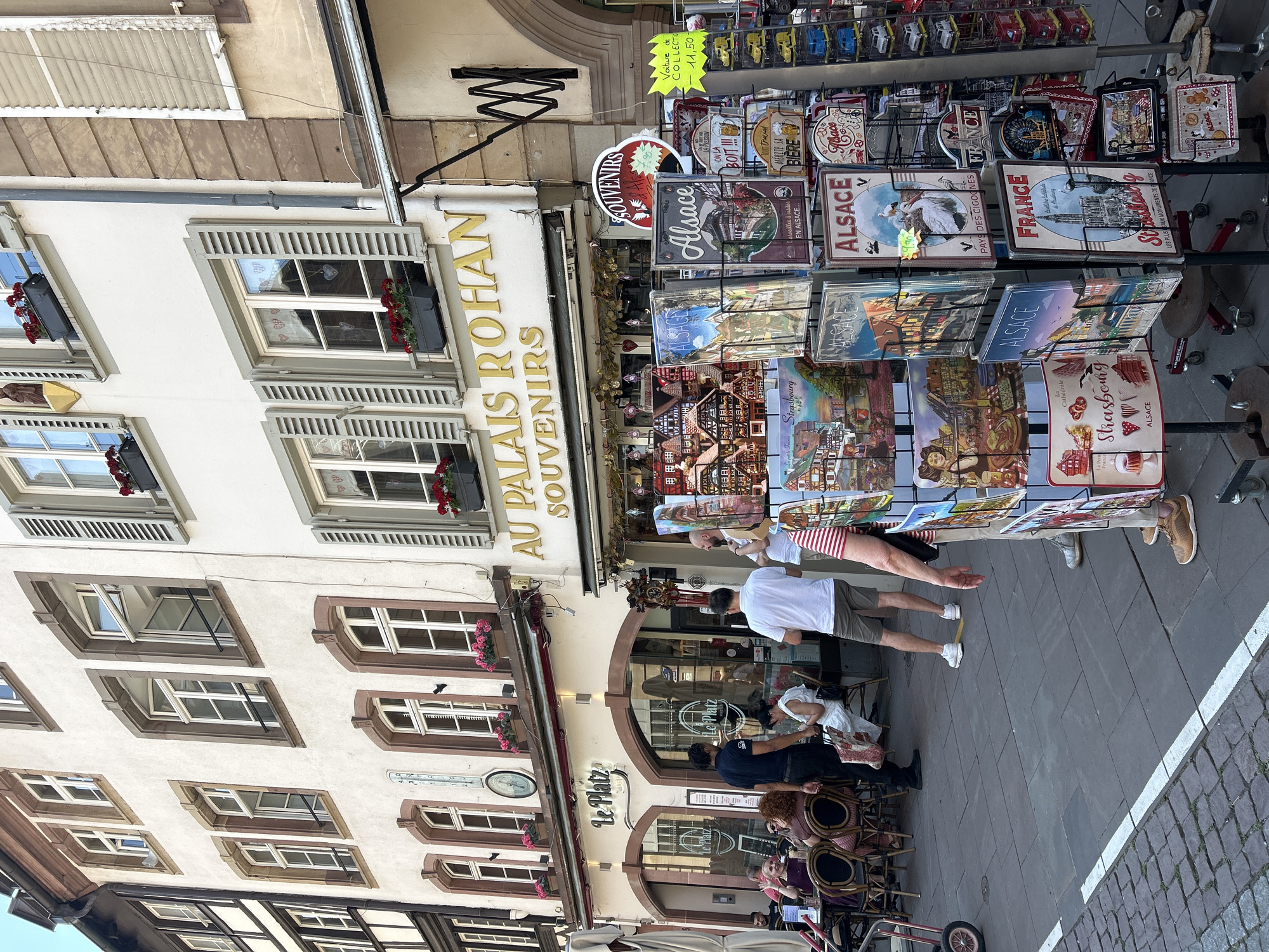 The image shows a street scene in Strasbourg, France, focusing on the exterior of shops and pedestrians. In the foreground, a display rack is filled with numerous souvenirs, primarily featuring images of Alsace and Strasbourg landmarks, such as the Strasbourg Cathedral. These souvenirs include metal signs, postcards, and miniature models of Alsatian houses. To the left of the souvenir rack, a restaurant or cafe called "Le Platz" is visible, with several patrons seated at outdoor tables. A waiter in a black t-shirt is interacting with customers. Further to the left, an awning shades more outdoor seating.
The buildings in the background are typical of European architecture, with multi-story structures featuring various window styles and decorative elements. One building prominently displays a sign reading "AU PALAIS ROHAN SOUVENIRS" in large gold lettering, indicating a souvenir shop. Another building to the left has a distinctive half-timbered facade.
People are casually dressed and engaged in various activities, such as dining, shopping, and walking. The street is paved with cobblestones, and there is a white line painted on the pavement in the lower left corner. The overall atmosphere suggests a bustling tourist area on a bright day.