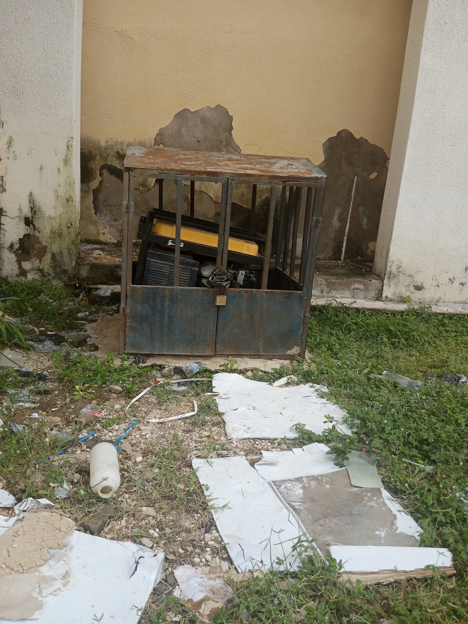 The image depicts a generator enclosed in a rusty metal cage, situated in an outdoor setting. The cage is positioned in front of a deteriorating building with a yellowish, textured wall showing patches of peeling paint and dark staining, likely from water damage or mold. To the left of the cage is a cream-colored wall, also showing signs of wear and discoloration.
The ground around the cage is littered with debris, including broken pieces of white material (possibly plaster or tiles), plastic bottles, and dry vegetation. Green grass and weeds are growing throughout the area, suggesting an unkempt or abandoned space. A white plastic bottle lies on the ground near the foreground, and a length of white tape or ribbon is also visible.
The generator inside the cage appears to be a portable model with a yellow fuel tank and black casing. The cage has a metal door with a padlock, indicating an attempt to secure the generator. Behind the cage, stairs can be seen leading up to a darker area, possibly an entrance to the building.
The overall impression is one of neglect and disuse. The time of day is not clearly indicated, but the lighting suggests it is daytime. The weather appears overcast or at least not intensely sunny, given the muted tones and lack of harsh shadows. The location cues, such as the building's architecture and the surrounding vegetation, combined with the provided location context of Jalingo, Nigeria, suggest a tropical or subtropical environment. There is no visible text in the image.