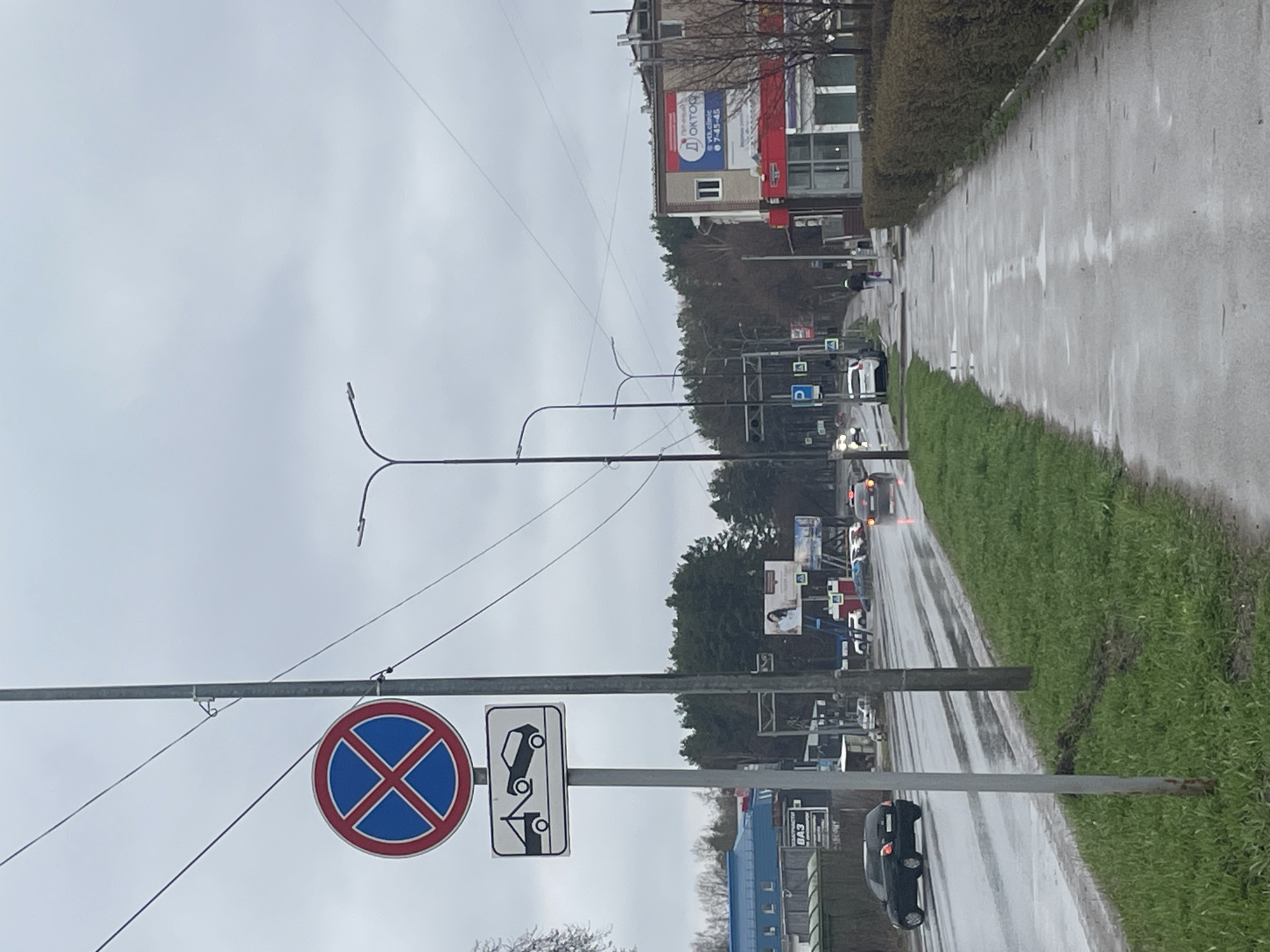 This image captures an overcast daytime street scene in Chaikovsky, Russia, likely after a period of rain, as evidenced by the wet asphalt and grass. The perspective is from the side of a road, looking down its length.
In the immediate foreground, a tall pole supports two prominent traffic signs. The first is a circular "No Stopping" sign (a blue circle with a red cross), and below it, a rectangular supplementary sign depicting a car being towed by a tow truck, indicating a "Tow Away Zone."
The road appears to have at least two lanes or a wide shoulder, with several vehicles visible. Some cars have their headlights or taillights on. A person is seen walking on a distant sidewalk on the opposite side of the street. Streetlights with distinctive double-curved arms line the road. A strip of green, wet grass separates the roadway from a sidewalk or verge on the right side of the frame.
Along the far side of the street, a line of trees partially obscures buildings behind them. One building prominently displays a red and blue banner with Cyrillic text that reads "Д ОКТОР" (D OKTOR), likely for a medical facility or pharmacy, along with a phone number "7-43-43" and the word "ВЫХОДНОЙ" (WEEKEND). Further down the road, another billboard and a partially visible gas station sign with "БАЗ" (BAZ) are present. The sky is uniformly light grey and cloudy.