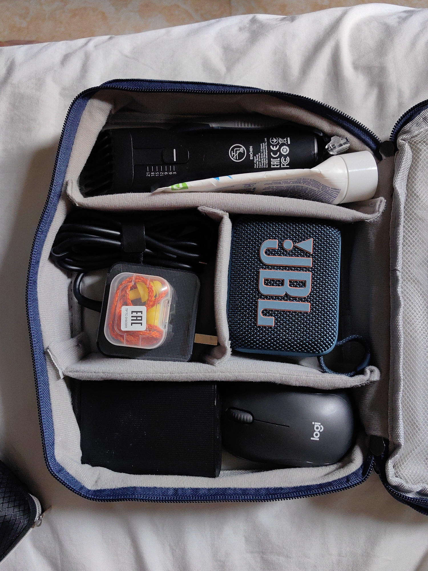 The image shows a partially opened, padded organizer bag, filled with various items. At the top, there's a black ENCHEN hair clipper resting next to a white tube of cream or lotion. Below that, there's a compartment containing a coiled black cable secured with a velcro strap, along with a small, clear plastic container holding orange earplugs, resting on a black power adapter. Next to that compartment is a blue JBL Bluetooth speaker with a gray cord. In the bottom compartment, there's a black power bank beside a black Logitech wireless mouse. The bag has a gray interior and a blue zipper. The bag is lying on a white surface that appears to be bedding.