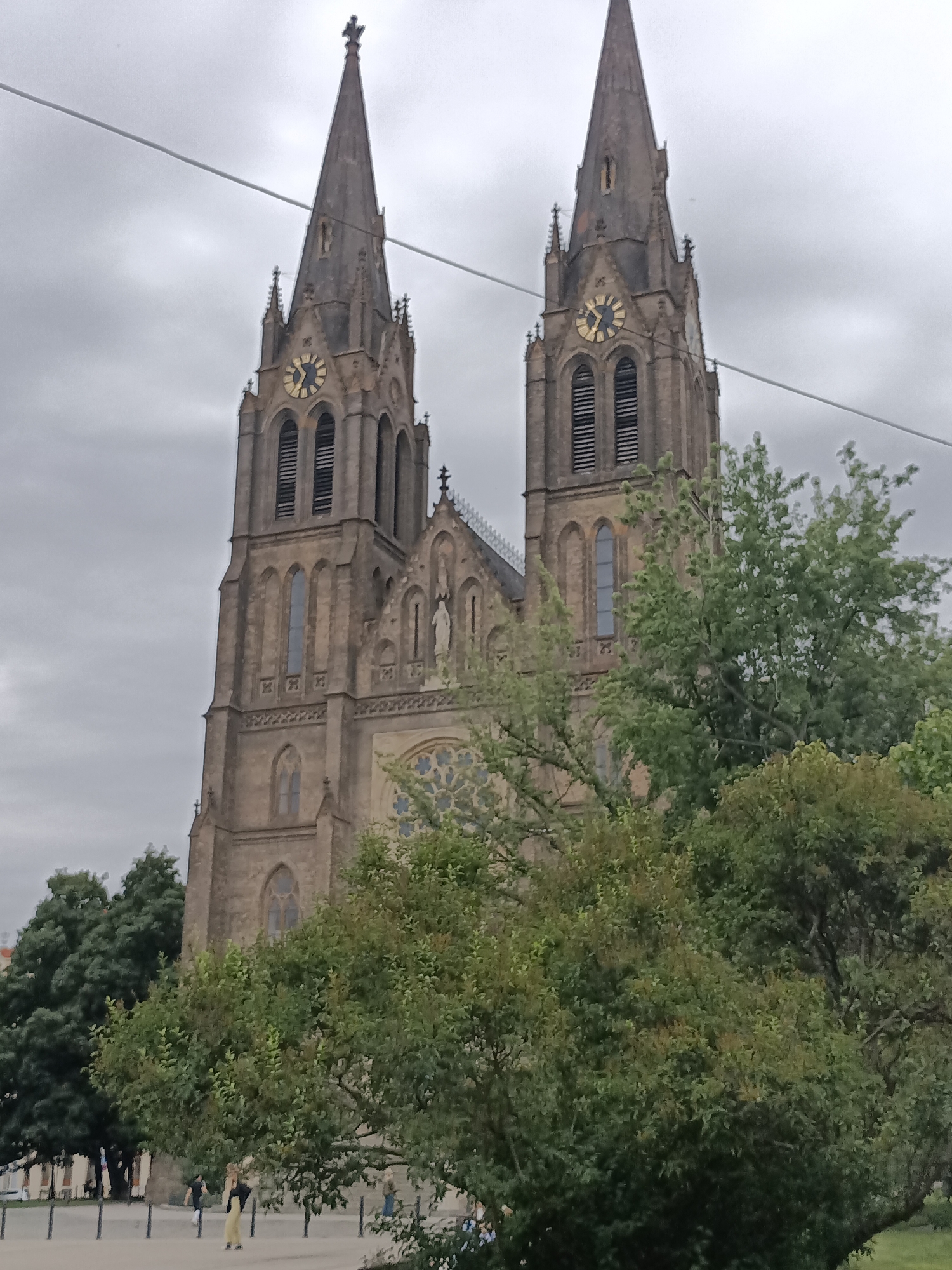 The image shows the Saint Vitus Cathedral, a prominent landmark located within Prague Castle in Prague, Czechia. The cathedral is characterized by its two tall spires, one of which features a clock face. The sky is overcast, and the lower portion of the image is obscured by trees. In the foreground, several individuals are visible walking on a paved area. A thin wire runs horizontally across the upper part of the frame.