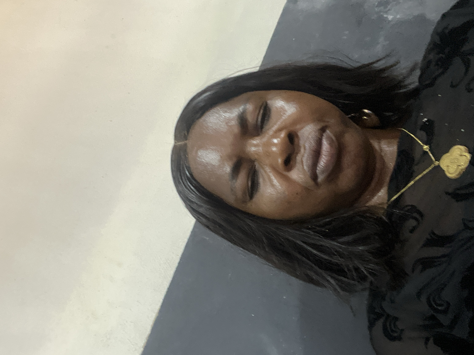 A close-up, slightly angled portrait shows a dark-skinned woman with closed eyes and dark hair. She is wearing a dark, patterned garment and a gold necklace with a pendant. The background is a two-toned wall, with the upper portion appearing light and the lower portion appearing dark gray. No further details indicate the location is Ado, Nigeria.