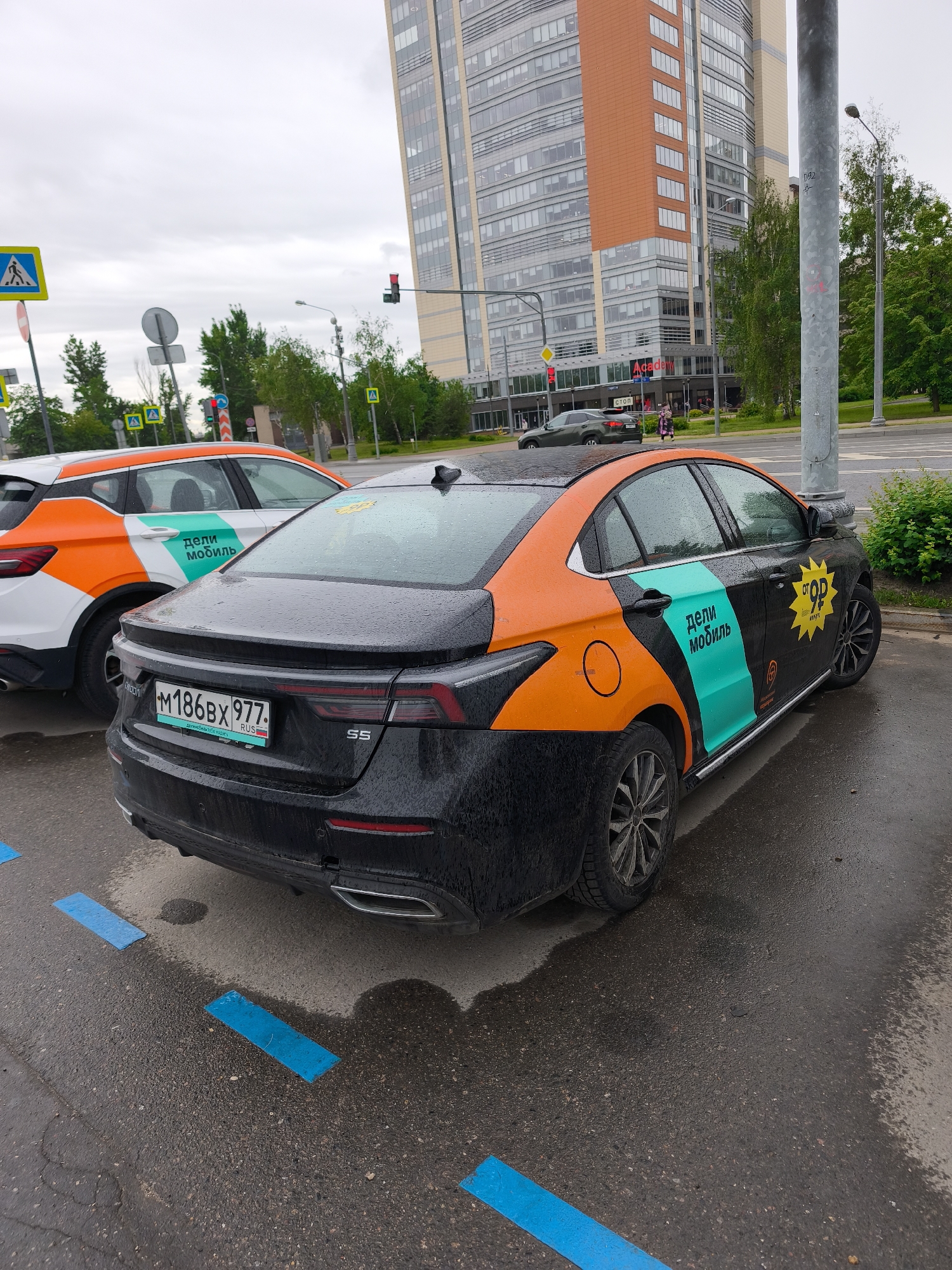 This is an exterior shot of a street scene in Moscow, Russia, likely taken on a cloudy day. Two cars are parked in blue parking spaces. The car in the foreground is a black sedan with orange and turquoise accents, including the words "дели мобиль" in Cyrillic. The license plate number "M186BX 977" is visible. The car next to it is white with orange and turquoise markings. Street signs and pedestrian crossings are visible along the road. In the background is a modern high-rise building. The overall scene suggests a typical urban environment.