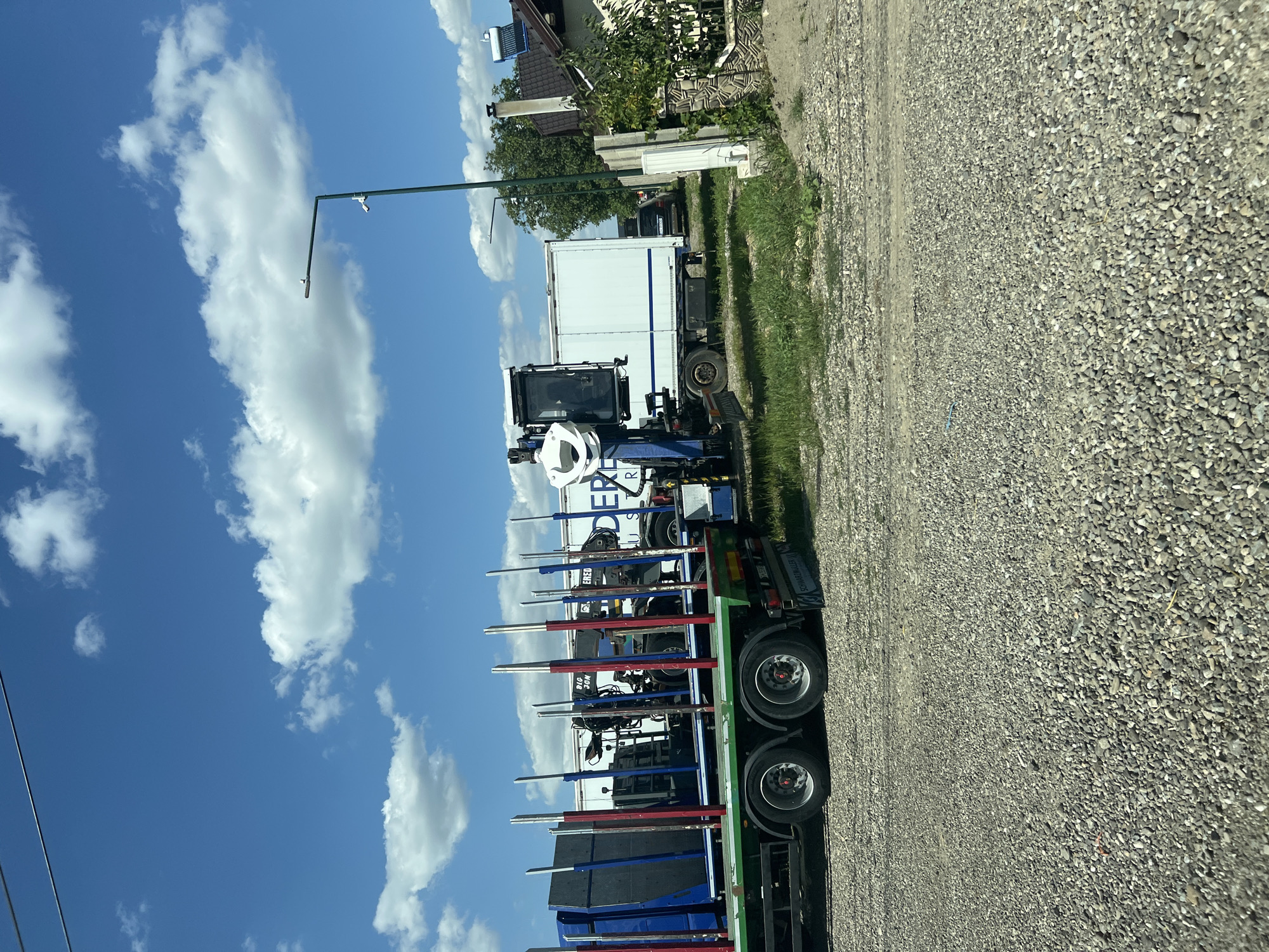 In Sasca Nouă, Romania, a sunny day with scattered clouds is depicted. The foreground is a gravel-covered ground. Two trucks are parked side-by-side on a patch of grass and gravel. The truck on the left is a flatbed trailer with tall, vertical poles, some red and some blue, along its sides, and what appear to be large, flat blue objects secured within. The truck on the right is a box truck with a crane attached to its side. The crane has a white cab and a large white grapple. The backdrop includes a green fence, some trees, and a building with a solar panel on its roof. A street light stands near the trucks.