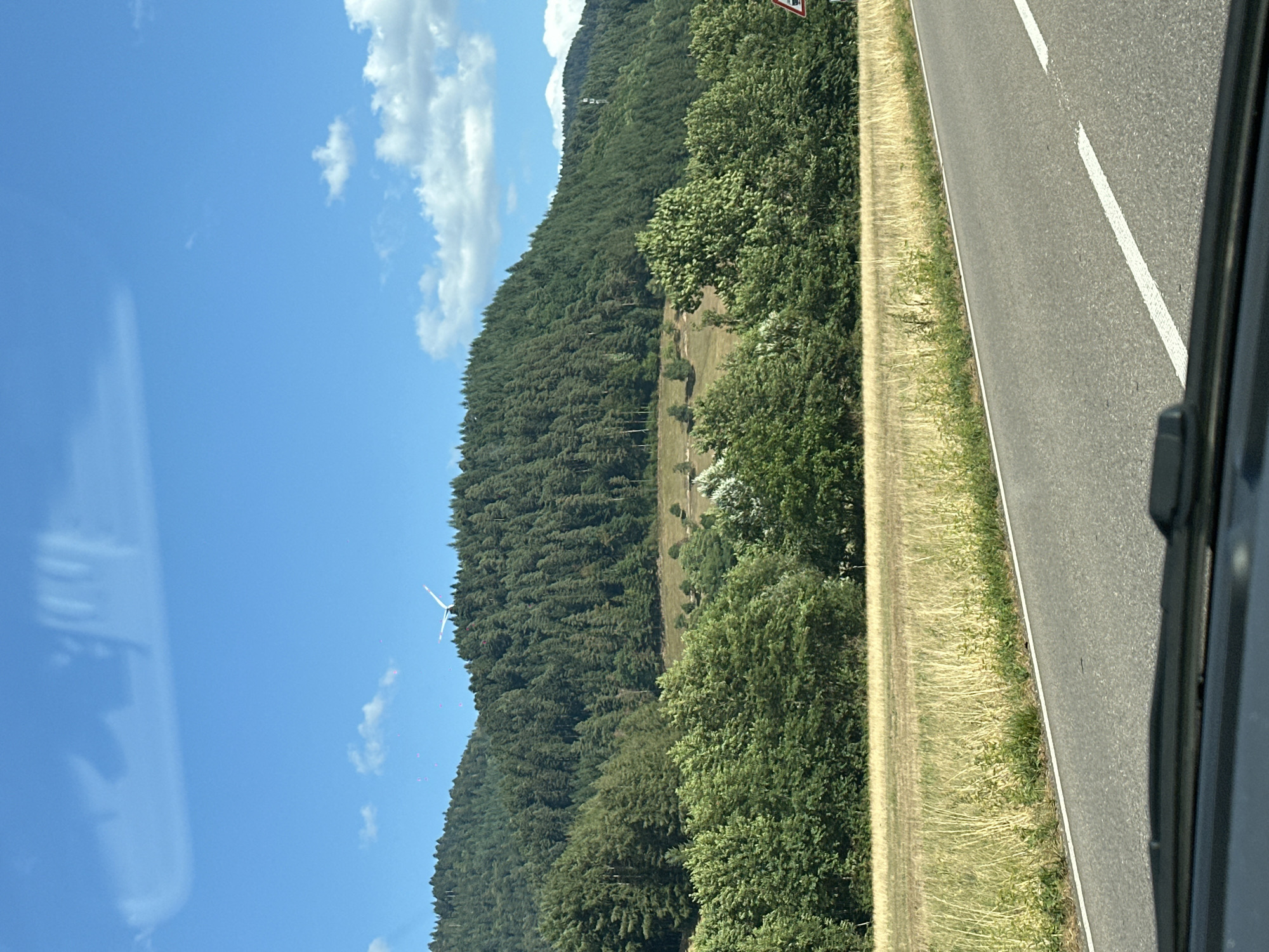 The image shows a scenic view of a rural area with rolling hills covered in dense green forest. A clear blue sky with scattered white clouds dominates the upper portion of the frame. In the distance, atop one of the forested hills, a white wind turbine is visible. In the foreground, a paved road runs horizontally across the image, bordered by a dry, grassy field. The perspective suggests the photo was taken from a moving vehicle, as the lower part of the image shows a portion of a car's dashboard. There are no people or animals visible in the scene. The landscape is characteristic of the Black Forest region, likely in or near Gutach (Schwarzwaldbahn), Germany, given the presence of forested hills and a wind turbine, common features in this area.