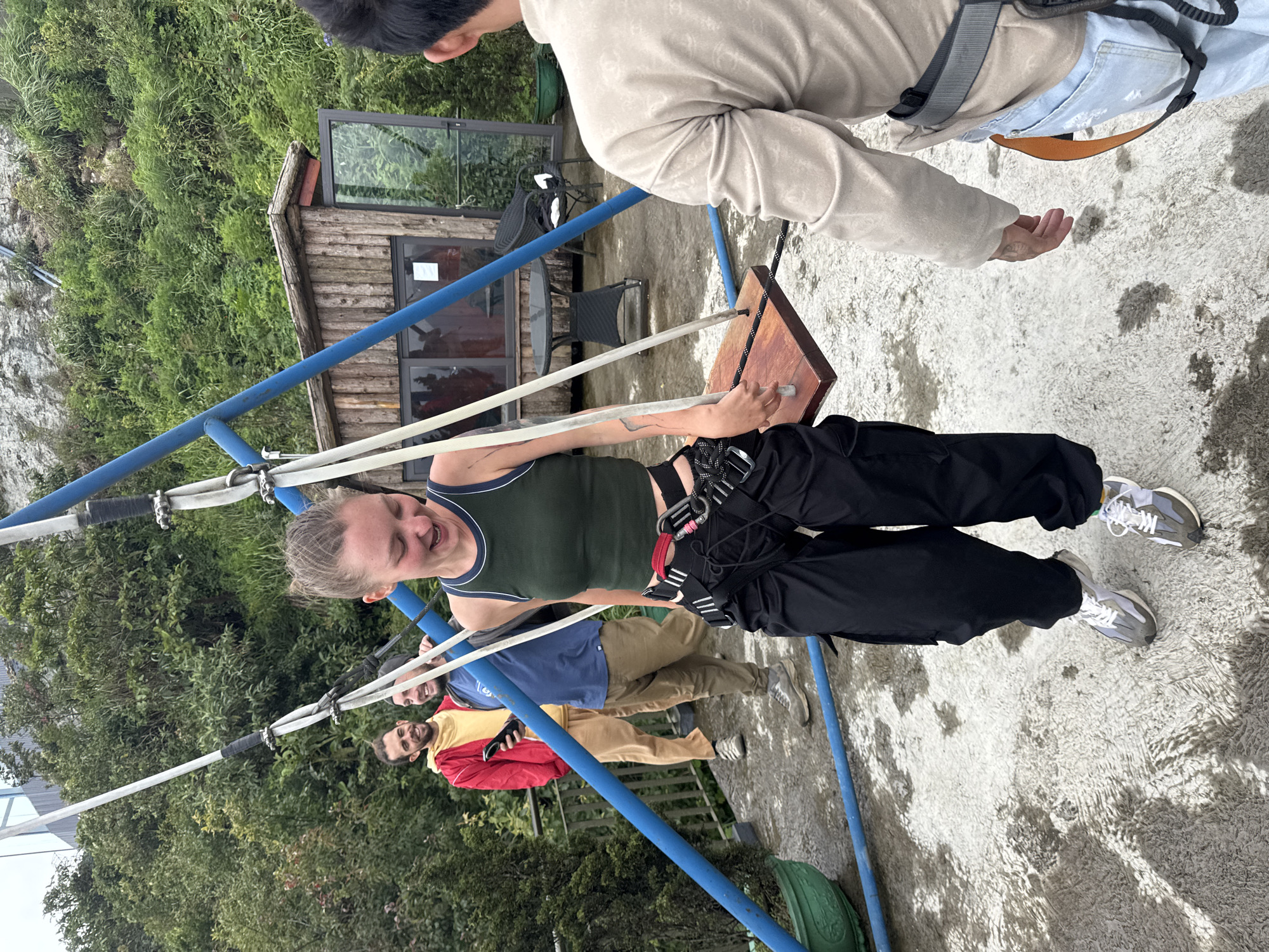 A woman in a green tank top and black pants is swinging on a wooden swing attached to a blue metal frame. She has a safety harness on. Behind her, two men stand on a platform; one is wearing a red and yellow jacket, and the other is wearing a blue shirt. Above her, another person in a beige jacket is extending a hand towards the swing. A wooden structure with windows is visible behind the swing set, along with lush green vegetation. The ground appears to be a concrete surface. This scene takes place in Binh Lu, Vietnam.