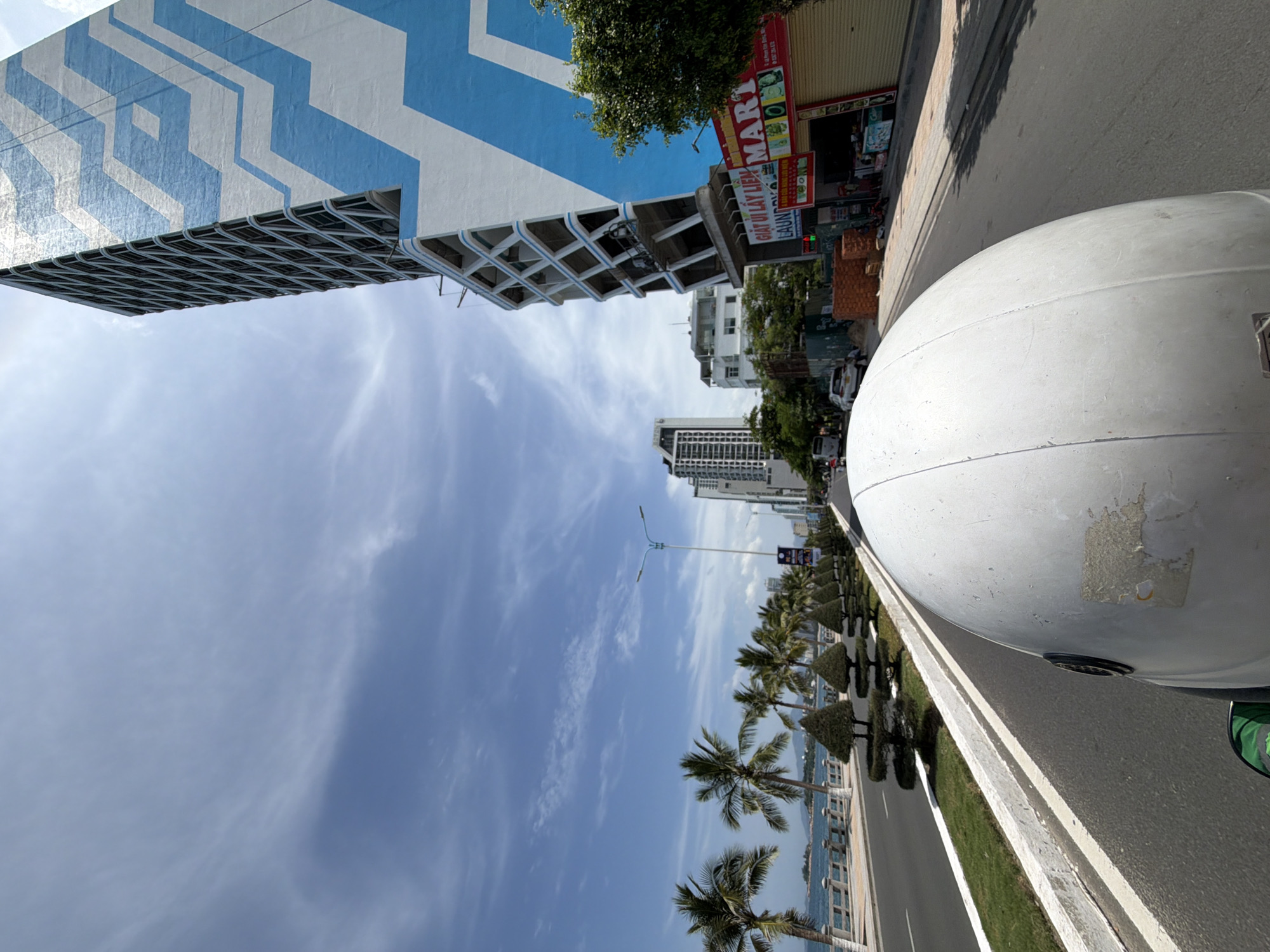 The image shows a view from a motorcycle in Nha Trang, Vietnam, looking down a street lined with palm trees and buildings. In the foreground, the white helmet of the rider is visible. The street is paved and has white lane markings. To the left, there is a sidewalk and a body of water, likely the sea, with a stone wall and railing. Beyond the water, there are distant islands. The right side of the street features several buildings, including a tall skyscraper with a blue and white zigzag pattern, and a smaller building with shops at street level, one of which is labeled "MART" and another with Vietnamese text. There are cars and a bus visible on the street, and pedestrians in the distance. The sky is blue with scattered clouds. The image appears to have been taken during daylight hours.