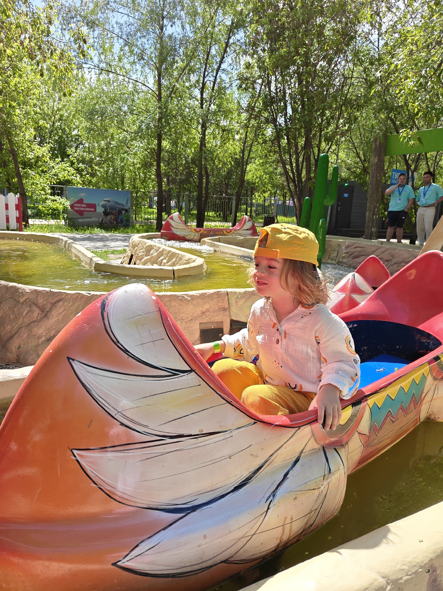 A fair-skinned child with blondish hair wearing a yellow baseball cap, a white button-up shirt with colorful designs, and yellow pants is seated in a boat-like structure within an amusement park ride. The boat has a red and orange exterior with painted designs. The ride appears to be water-based with multiple boats in a channel of light green water with concrete bordering. In the background, there is greenery, trees, and structures made to look like cacti. Further in the background, two adult males are standing and watching. A sign is visible in the far background. The location is Khimki, Russia.