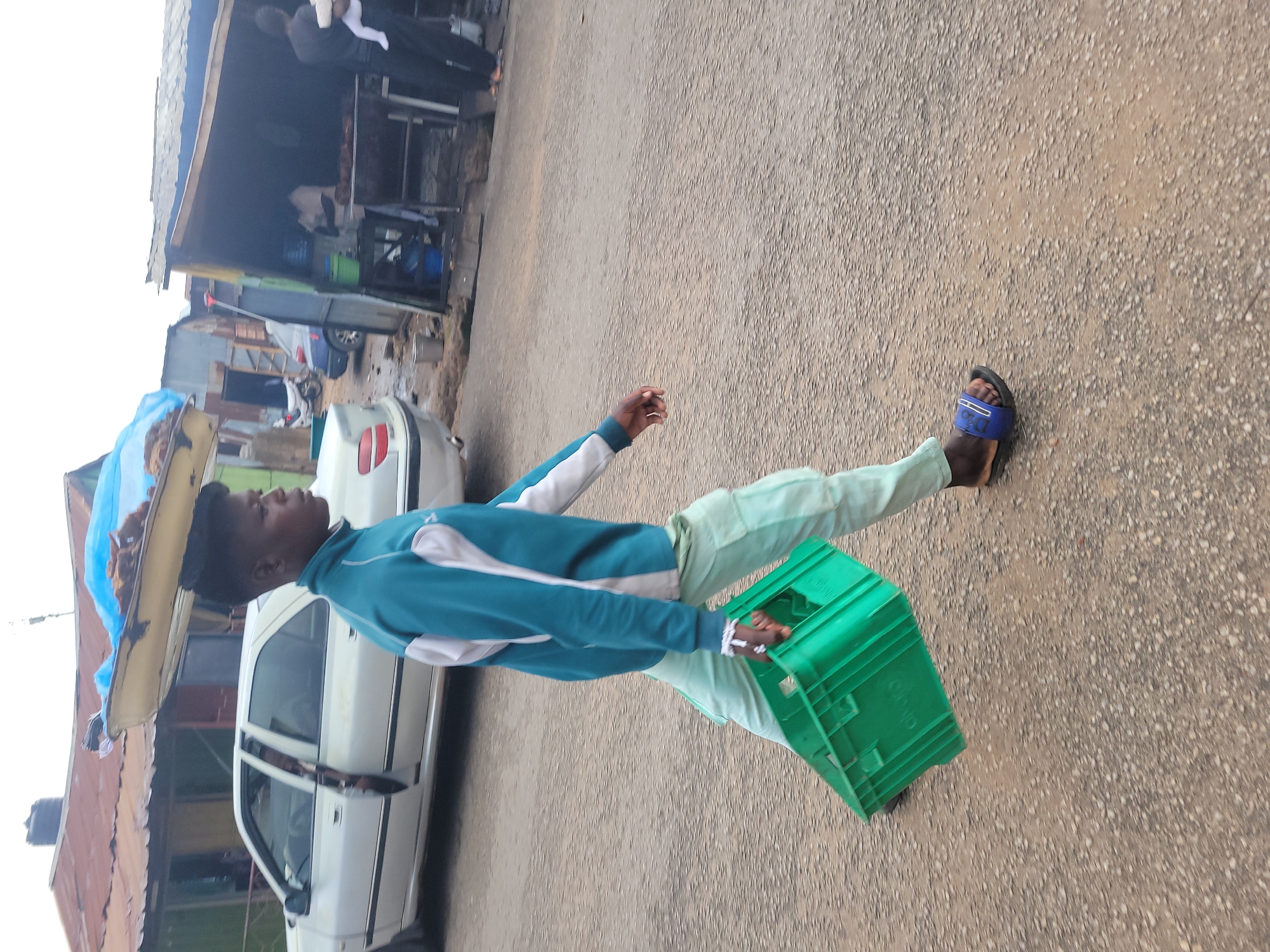 A person is walking with a crate in their left hand and a tray of goods balanced on their head. The person is wearing a teal and white tracksuit and light green cargo pants, with blue and white slides on their feet. The crate is green and rectangular. The tray on their head contains what appears to be dried fish covered by a blue tarp. The person is walking on a gravelly road in front of a row of small businesses. In the background, a white sedan is parked, and a few other structures and vehicles are visible. The scene is likely in Jos, Nigeria, given the presence of this type of open-air market and transport of goods.