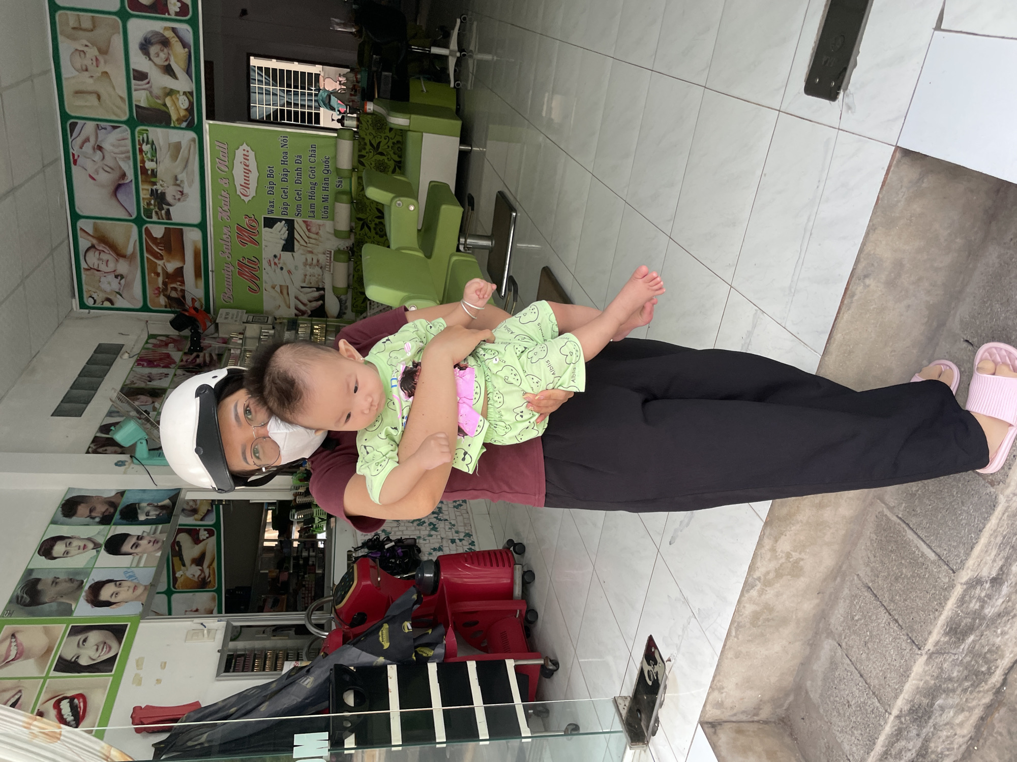 A woman wearing a white helmet and a face mask holds a baby in front of a beauty salon in Phu Thuy Ward, Vietnam. The woman is wearing a maroon t-shirt and black pants. The baby is dressed in a light green outfit with cartoon characters. The salon appears to be open, with clients visible in the background. The floor is tiled, and there is a step leading up to the salon entrance. The overall scene suggests a typical day at a local business.
