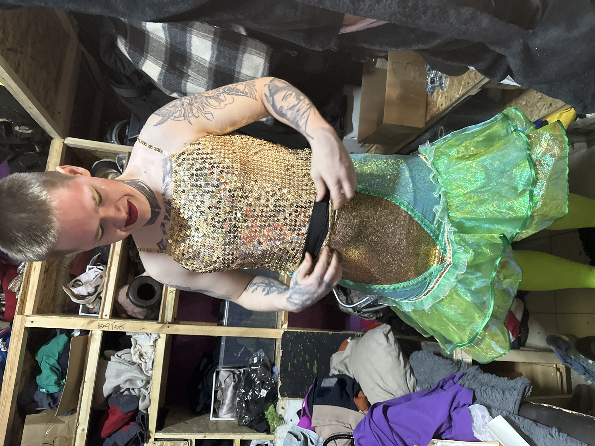 An adult individual with short, light-colored hair, wearing elaborate makeup including red lipstick, yellow eyeshadow, and glitter, stands indoors, looking downwards at their attire. A nose piercing is visible, along with extensive tattoos on both arms and the neck, including a black band-style tattoo. The individual wears a sleeveless, gold sequined top with thin straps, a metallic gold and teal/green skirt featuring a ruffled, translucent green overlay, and bright green leggings. Their hands are positioned at the waist of the skirt, appearing to adjust the garment.
The setting is an interior space with a cluttered background. To the left and behind the individual, a multi-tiered wooden shelving unit, constructed from unfinished planks, is packed with various items. These include folded clothes in different colors (green, grey, red, black), fabrics, and brown cardboard boxes, one of which has "B3" handwritten on it. To the right of the individual, dark fabrics, including a plaid blanket or garment, hang from an unseen fixture. The floor, partially visible, appears to be a light-colored, possibly tiled or concrete surface, also showing some clutter. The environment suggests a backstage area, a costume storage room, or a workshop.
