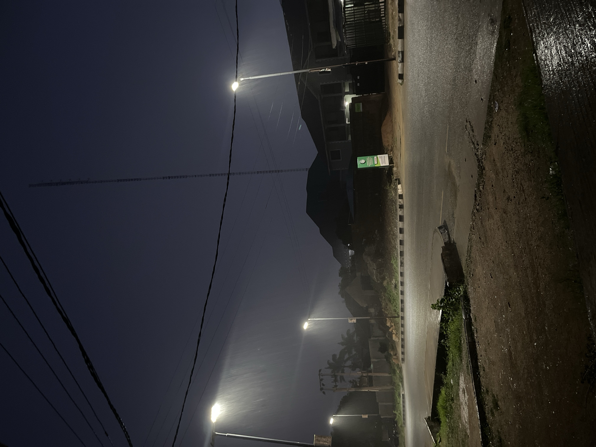 The image depicts a street scene at night during heavy rainfall in the city of Jos, Nigeria. Three streetlights illuminate the road and sidewalk, their beams creating visible streaks in the downpour. The road surface is wet and reflective, with puddles forming in places. On the right side of the image, there is a building with a dark facade and a metal gate. To the left, another building is visible, partially obscured by trees. A tall radio tower stands prominently in the background against the dark sky. Power lines are strung across the upper portion of the frame. The overall atmosphere is dark and damp, characteristic of a rainy night.