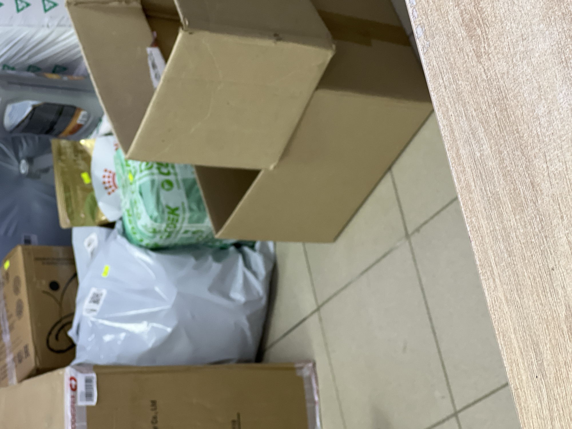 The image depicts a cluttered indoor space, likely a storage area or a shop, with various cardboard boxes and packages. In the foreground, a wooden surface is visible, possibly a table or counter. The background is filled with stacked boxes of different sizes and materials. Some boxes are plain cardboard, while others are branded or have labels. A grey plastic-wrapped package is prominent, and a green package with text is also visible. A black plastic bottle, possibly motor oil, is situated behind some of the boxes. The floor appears to be tiled. The overall impression is one of disorganization and storage. There are no people visible in the image. The location is identified as Unknown, Russia.