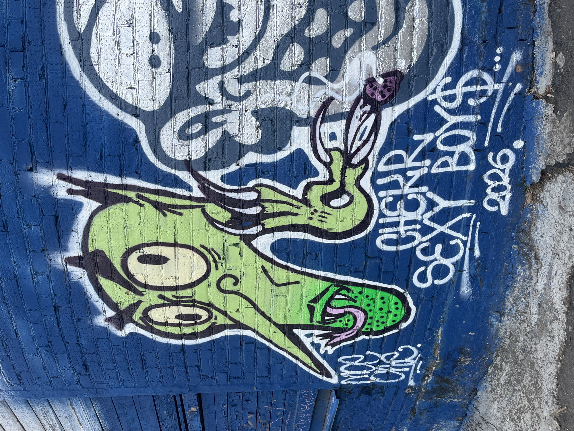A close-up shot shows graffiti on a blue brick wall in Krasnoyarsk, Russia. The graffiti consists of a cartoon-like green monster with large, bulging eyes, a wide open mouth revealing a green tongue with dark green polka dots and a pink end. The monster appears to be smoking a joint with purple dots on its tip. Above the monster is a cloud of white smoke. To the right of the monster, white spray-painted words read "SEX $ CHLDRN BOY $" and "2026". Below the monster, there are two overlapping, illegible tags in white spray paint. The brick wall is painted a deep blue color, except for a small portion on the right where the brick is exposed and painted grey. The lower part of the image includes what appears to be a portion of blue painted wood.