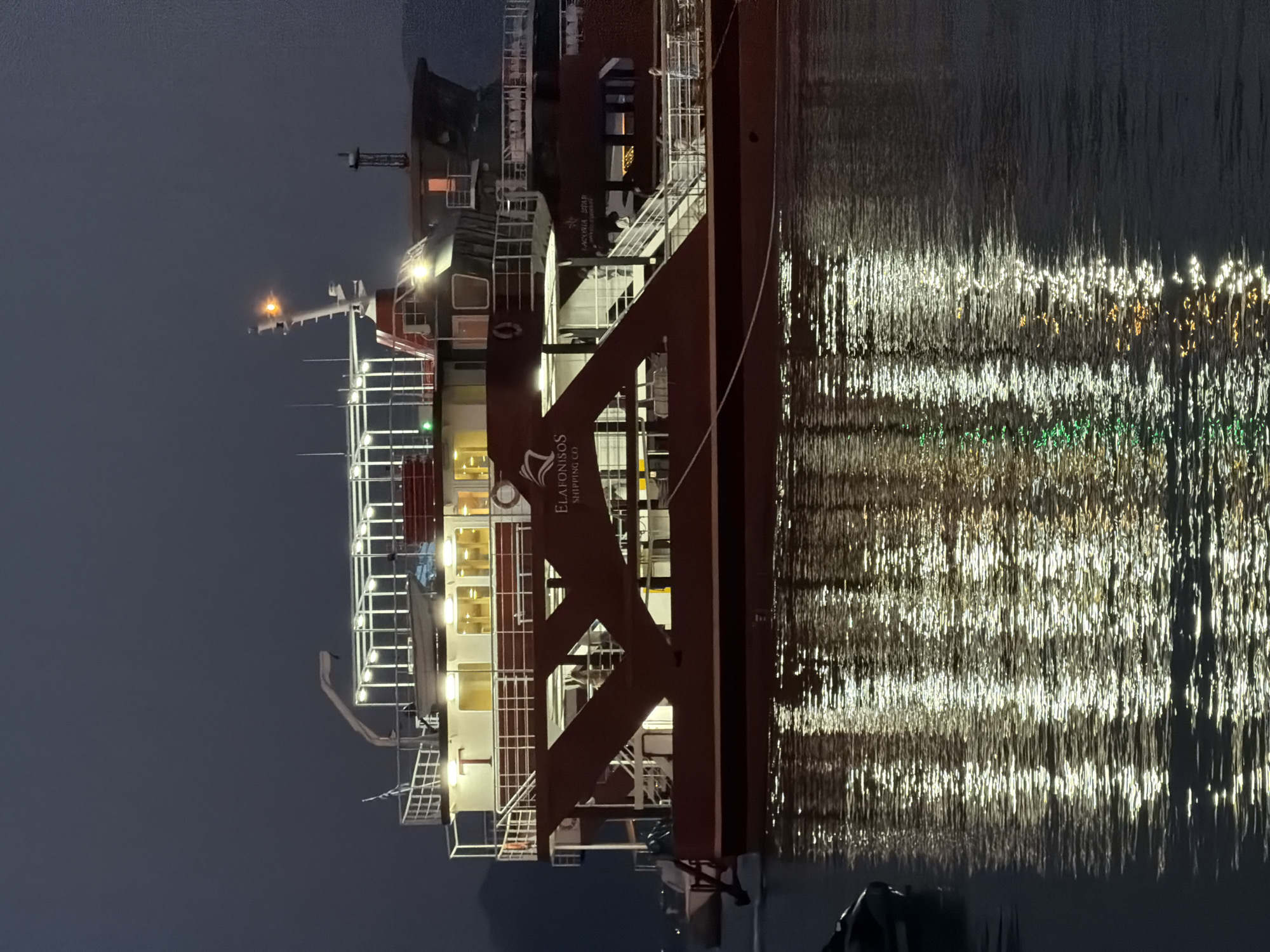 A large, maroon and white ferry, named "Elafonisos Shipping Co.", is docked at night in the harbor of Elafonisos, Greece. The ferry is brightly illuminated by interior and exterior lights, reflecting on the dark, rippling water. The ferry has multiple decks with railings and windows, and a crane is visible on the upper deck. A small inflatable boat is tied to the side of the ferry, partially submerged in the water. The sky is dark and overcast, with faint lights visible in the distance.