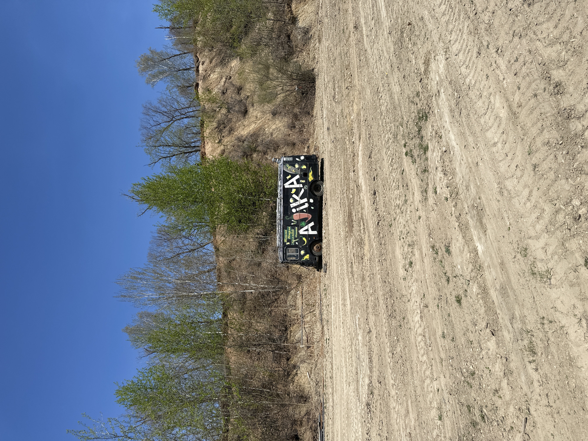 The scene is a dirt lot in Chigiri, Russia, with a black vehicle parked on the right side of the image. The vehicle has graffiti with the word "AIKKA" prominently displayed on its side. The lot shows tire tracks, indicating previous vehicular activity. Behind the vehicle is a steep, vegetation-covered slope. The sky is clear and blue.