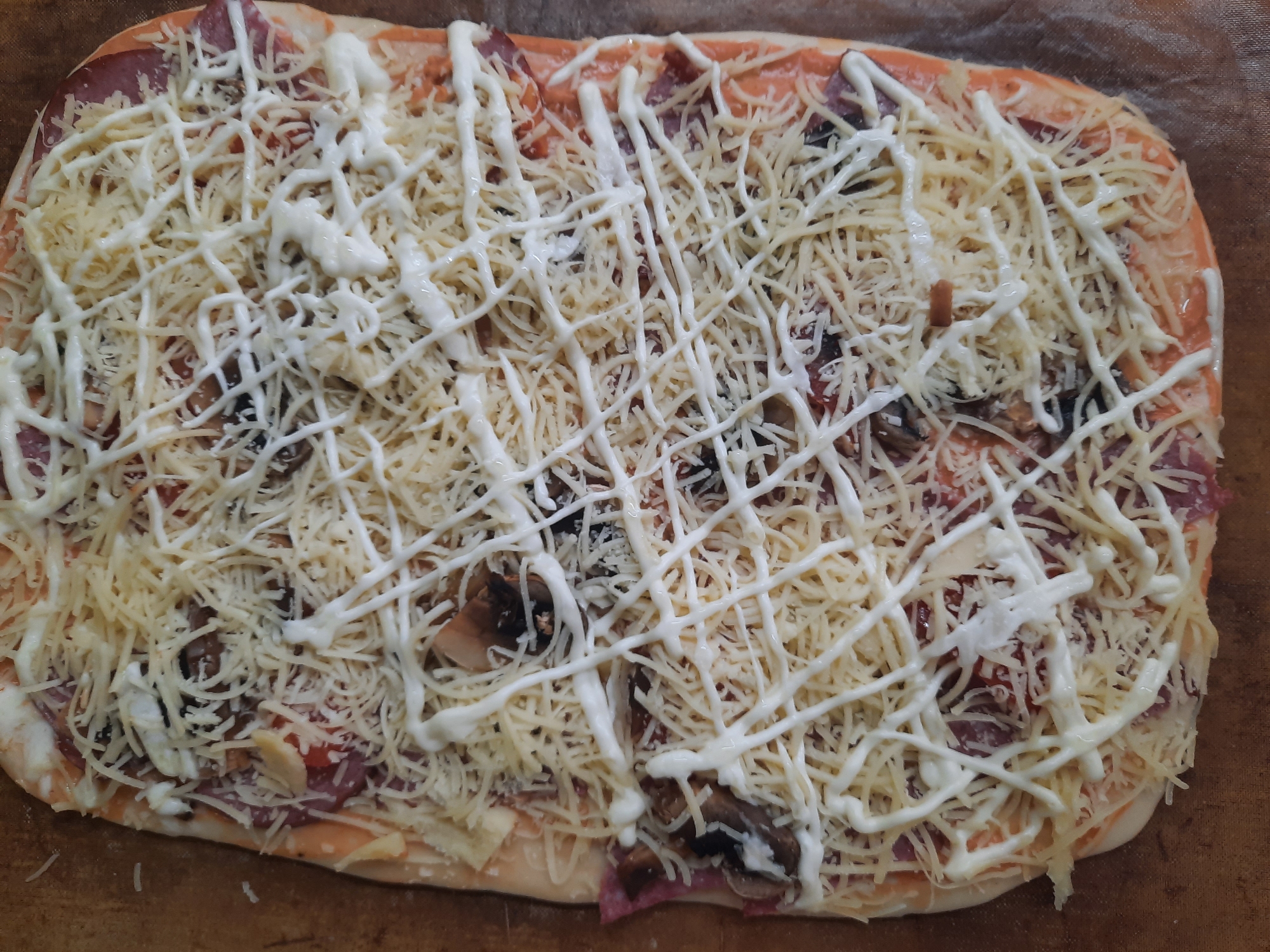 The image shows a rectangular pizza on a baking sheet. The pizza is topped with tomato sauce, salami, mushrooms, shredded cheese, and a drizzled white sauce, possibly mayonnaise or a similar condiment. The environment is indoor. The location is Unknown, Unknown.