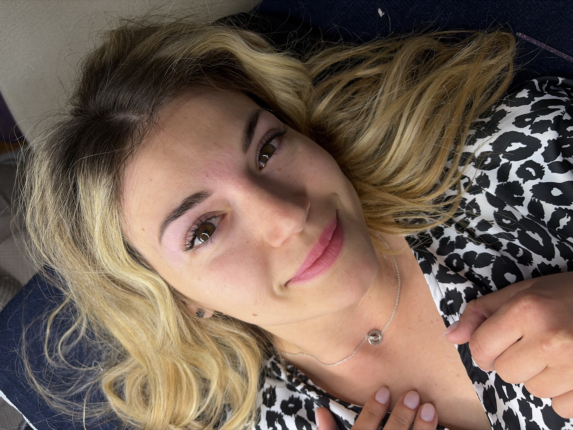 A woman is lying down, seemingly on a blue surface. Her hair is blonde, with darker roots visible. She is wearing a black and white patterned top and a silver necklace with a circular pendant. She is lightly smiling. The scene takes place in an indoor environment. There is no indication that this photo was taken in Budva, Montenegro.