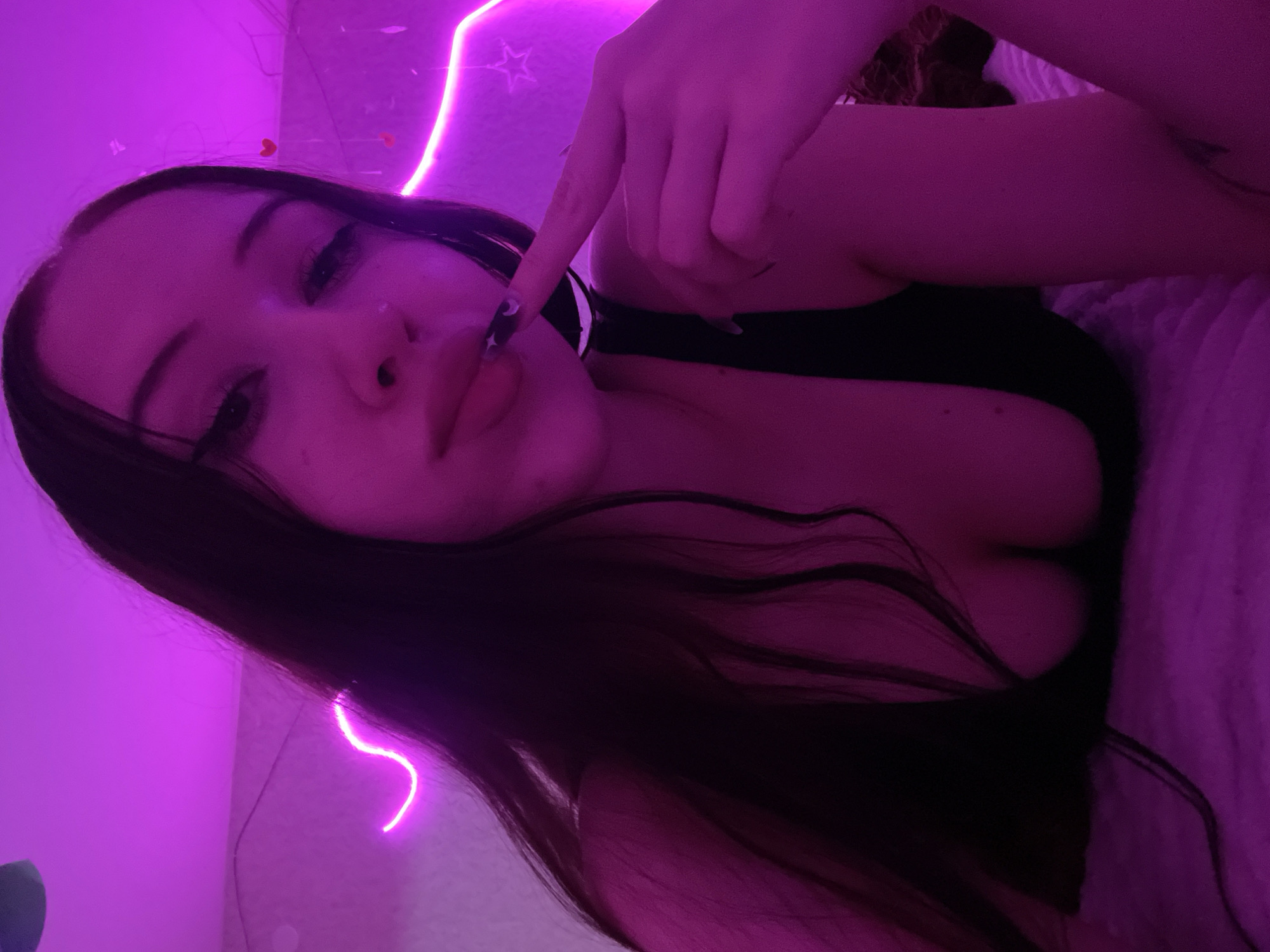 A young woman with long, dark hair is lying down, illuminated by pink neon lights. She is wearing a black top that reveals her cleavage and has long fingernails, one of which is touching her lips. The background is a wall decorated with pink heart shapes and a star. The scene appears to be indoors, possibly a bedroom. The specific location in Votkinsk, Russia, cannot be determined from the image.