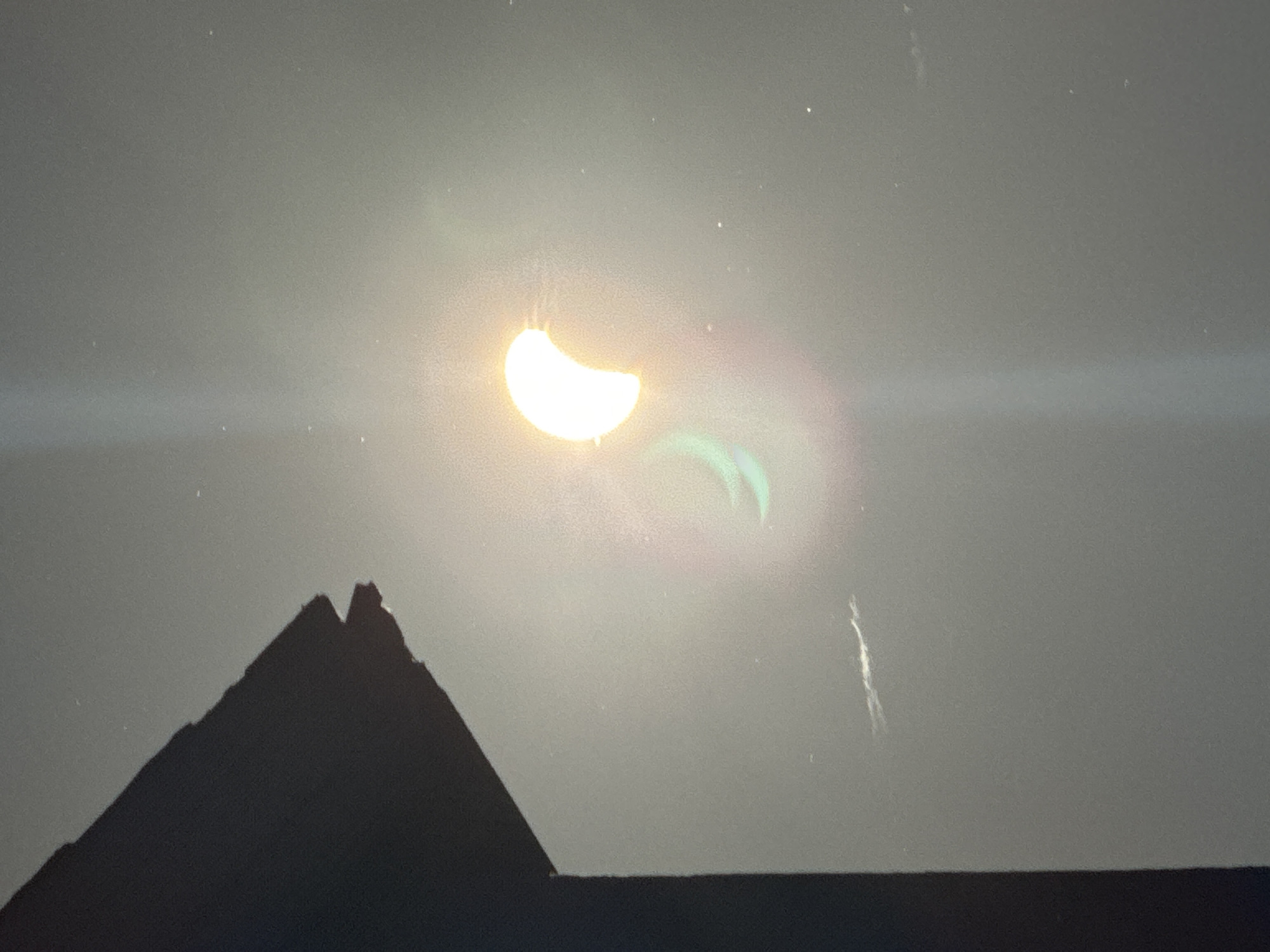 The image shows a partial solar eclipse viewed from Waterford, Ireland. A silhouetted structure, likely a building or roofline, is visible in the lower left quadrant, casting a shadow. The sun appears as a bright crescent shape, partially obscured by the moon. Lens flare creates a colorful halo effect around the sun, with green and pink hues visible. A thin, white, streaky object, possibly a contrail from an aircraft, is present in the sky. The overall environment appears to be daytime, with a hazy or overcast sky.