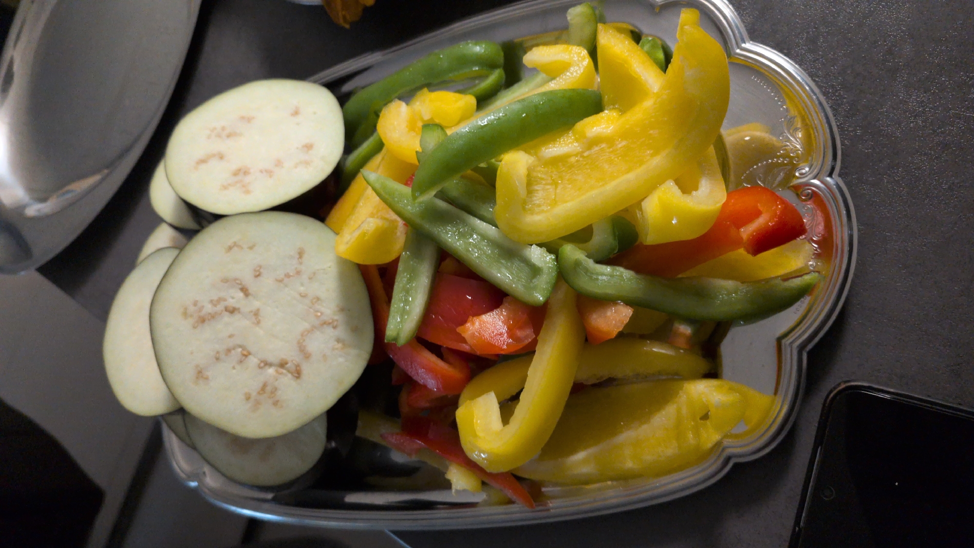 The image shows a close-up, overhead view of sliced eggplants and cut bell peppers arranged on a decorative silver platter. The eggplants are sliced into thick rounds and appear light in color with visible seeds. The bell peppers are chopped into strips and are a mix of yellow, green, and red. They are placed in the platter below the eggplant slices. The platter itself has an ornate, scalloped edge. In the bottom left corner of the image, the corner of a dark smartphone is visible, resting on a dark gray textured surface, likely a countertop. The background is out of focus, showing a hint of a darker surface and a metallic sheen from what appears to be another platter or surface. The scene is indoors and appears to be in a kitchen setting, potentially in Dissen am Teutoburger Wald, Germany, given the context provided. There are no people visible in the image.