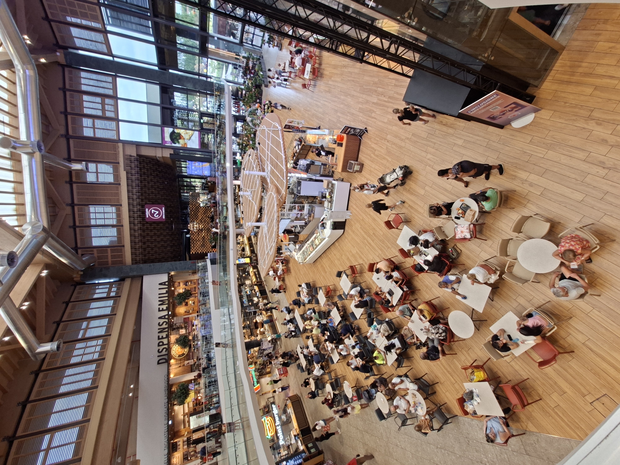 The image shows a high-angle, wide shot of the interior of the Il Centro shopping mall in Arese, Italy. The mall features a large, open-plan layout with multiple levels. The flooring is made of light-colored wood planks. The scene is populated with many people, most of whom are seated at tables in various food establishments or walking around.
In the foreground, there are several clusters of tables and chairs occupied by diners. To the left, a woman is seated at a table. Further to the right, a group of people are gathered around tables, some eating, some conversing. A stroller is visible near the center.
Further back, a food court area is visible with multiple vendors and seating areas. Signs for "DISPENSA EMILIA" and "Point Burger" are prominently displayed. People are ordering food, eating, and interacting.
The architecture of the mall is modern, with exposed metal beams and ductwork overhead. Large windows on the right side of the image offer a view of the outdoors, with greenery visible. A tall black metal structure, possibly an elevator or part of the mall's infrastructure, is also present. An advertising stand is positioned near this structure. The overall atmosphere appears to be busy and social, consistent with a shopping mall environment.