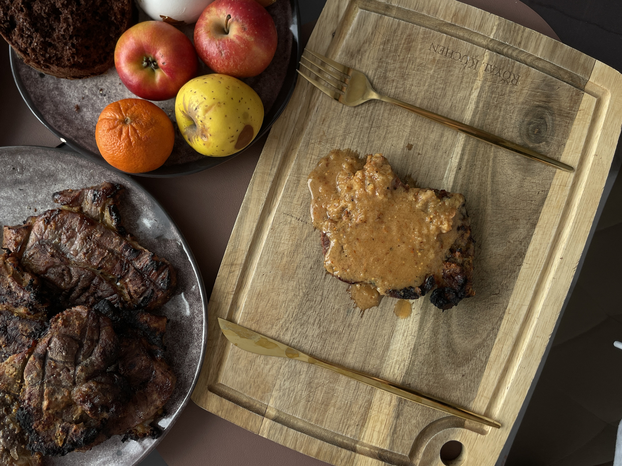 A high-angle shot captures a culinary arrangement on a neutral-toned surface, possibly a table, in what appears to be an indoor setting.
A slate-colored plate in the lower-left quadrant holds three pieces of what looks like grilled or pan-fried meat with visible charred sections. Adjacent to this plate is another slate-colored plate containing an assortment of food items. On this plate, from left to right, are a piece of dark bread, a white round object, three red apples, and a tangerine.
A wooden cutting board occupies the right half of the image. On the board rests a piece of cooked meat, possibly pork, covered in a beige-colored sauce. A gold-colored knife and fork lie parallel to the meat, one above and one below. The cutting board has an engraved inscription that reads "ROYAL KUCHEN."
No people are visible in the shot. The environment seems to be well-lit, though the specific location in Подшивалово, Russia cannot be definitively determined from the image alone.