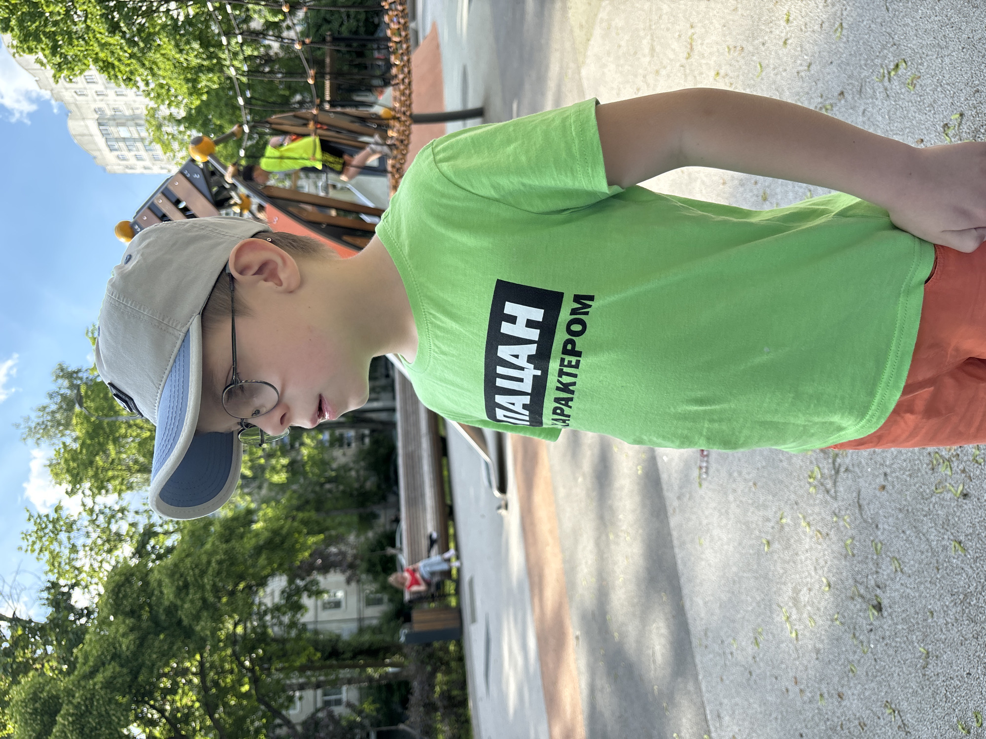 This image captures a young boy in an outdoor urban park or playground in Saint Petersburg, Russia, under bright daylight. The boy is positioned off-center, facing downwards and to his right, wearing a vibrant lime green t-shirt, orange shorts, a grey baseball cap with a light blue brim, and round-rimmed glasses.
The green t-shirt prominently displays Cyrillic text in a black box, reading "ПАЦАН С ХАРАКТЕРОМ," which translates to "A Boy With Character." His expression is neutral, suggesting focus or observation.
The background reveals a verdant environment with numerous trees, indicating a lush green space. A multi-story, light-colored building with multiple windows is visible behind the trees, characteristic of urban architecture. Playground equipment, featuring wooden and metal structures with yellow spherical elements, is partially visible. In the mid-ground, another person wearing a high-visibility yellow vest can be seen on the playground. Further in the background, a faint figure in a red top is seated on a bench.
The ground is a paved surface, likely asphalt or concrete, with scattered small green debris, possibly seeds or tiny leaves. The clear sky with some visible clouds suggests good weather, likely late morning or early afternoon. The overall scene depicts a casual, pleasant outdoor moment in a city park.
