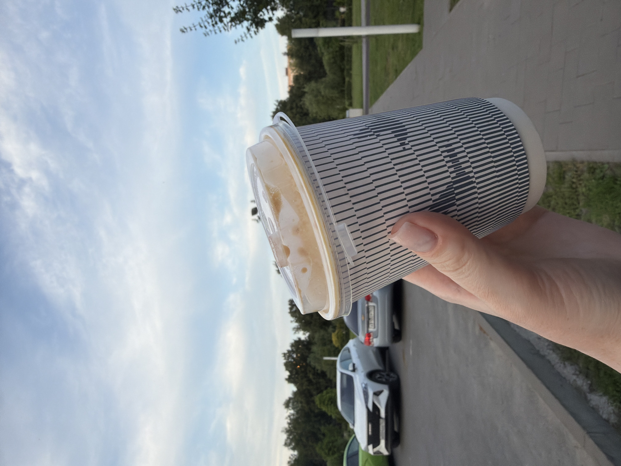 In Tolyatti, Russia, a hand holds a disposable coffee cup with a clear plastic lid, tilted towards a cloudy sky. The cup is covered in a black and white striped sleeve. In the background, a row of parked cars, including a white SUV and a silver sedan, are visible on a paved surface adjacent to a grassy area and trees. The scene appears to be an outdoor public space, possibly a parking lot or promenade.