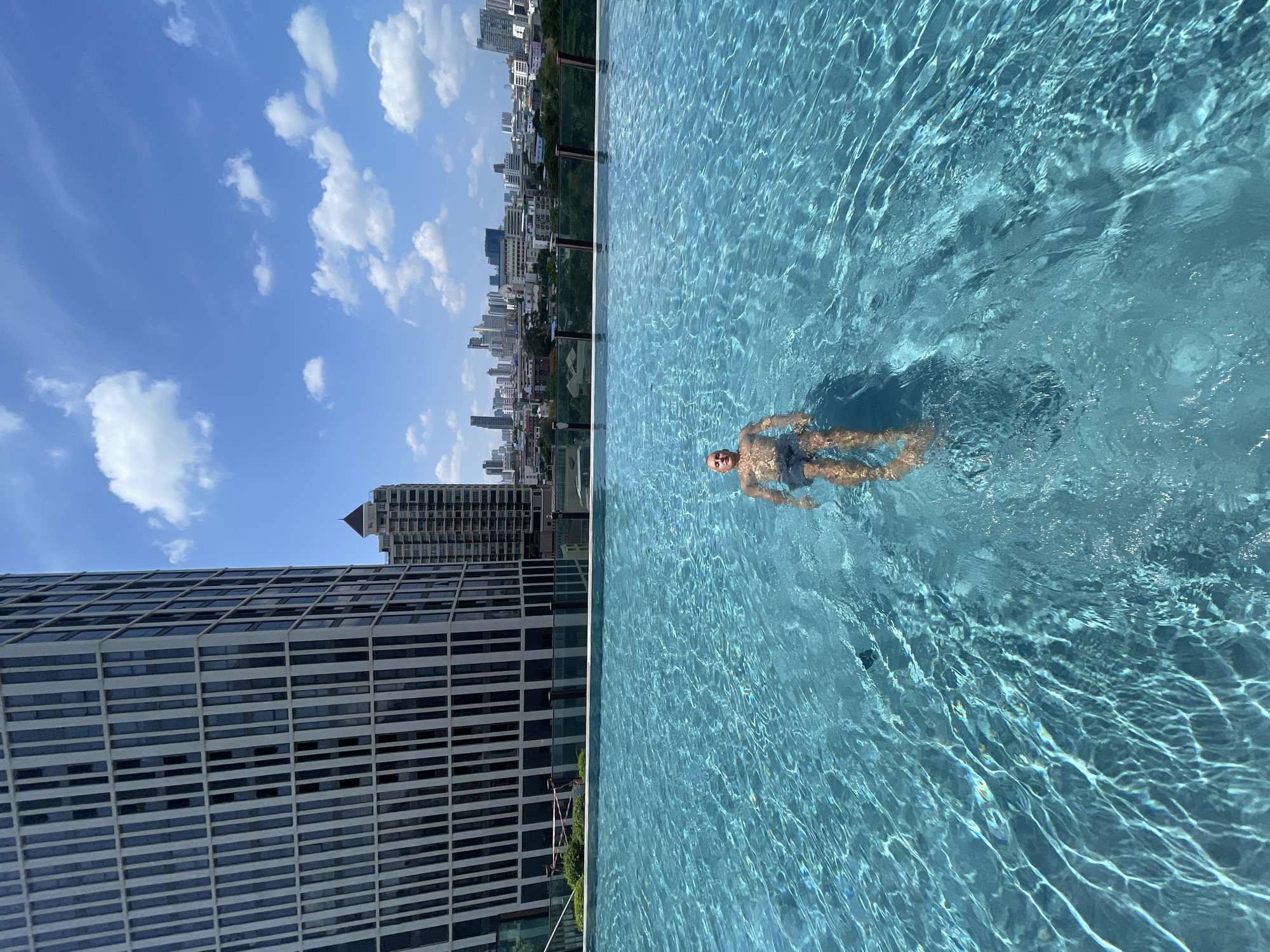 A man is swimming in a rooftop infinity pool on a sunny day. The water is clear blue with visible refractions. The man appears to be bald with light skin and is wearing dark-colored swimming trunks. The city skyline of Bangkok, Thailand, is visible behind the pool. The sky is blue with scattered white clouds. A tall modern building with a grid-like facade is on the lower left of the image.