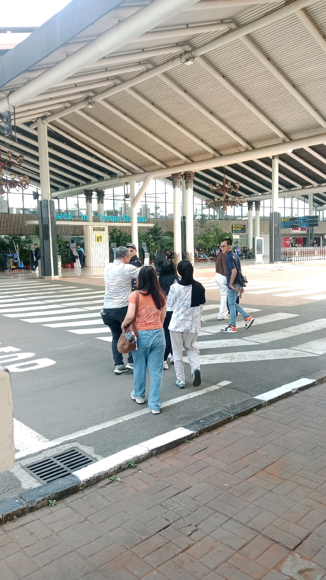 The image depicts a street scene outside an airport terminal in Tangerang, Indonesia. Several individuals are crossing a pedestrian zebra crossing. The foreground shows the legs of people walking, with one person wearing blue jeans and a light-colored patterned shirt, and another in light-colored pants and a dark headscarf. To the left, a person in light blue jeans and an orange t-shirt carries a brown bag. Behind them, a man in a white patterned shirt and dark pants is seen talking.
In the background, the airport terminal building is visible with its distinctive white, angled roof structure supported by pillars. Large windows and glass doors reveal the interior, where more people can be seen. The environment is a public space, likely an airport, with clear markings for pedestrian crossings and a paved sidewalk to the right. The lighting suggests it is daytime.