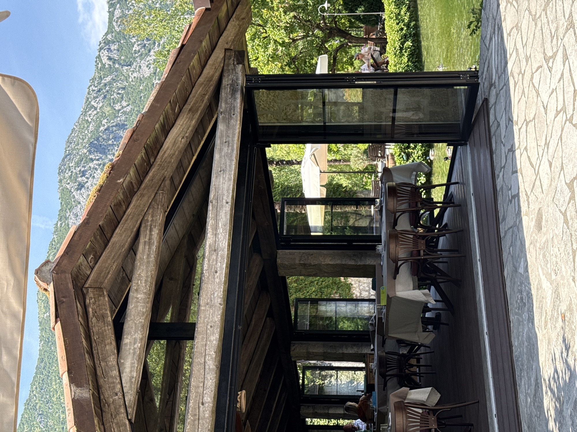 The image shows an outdoor restaurant seating area with tables covered in light-colored cloths. The seating area is covered by a wooden-beamed roof with terracotta tiles. In the background, there is a body of water, likely a lake or pond, with several ducks swimming. There are also trees and lush green foliage surrounding the area, with mountains visible in the distance under a clear blue sky. A person is seated at a table inside the covered area, and a couple is visible at a table in the background, on the right side of the image. The restaurant appears to be located in a scenic natural setting. The architecture of the covered seating area, with its rustic wooden beams and stone pillars, suggests a blend of traditional and modern design. The overall atmosphere is serene and inviting, suggesting a pleasant dining experience in a beautiful natural environment. This location is in Morinj, Montenegro, a picturesque village on the Bay of Kotor known for its natural beauty and tranquil atmosphere.